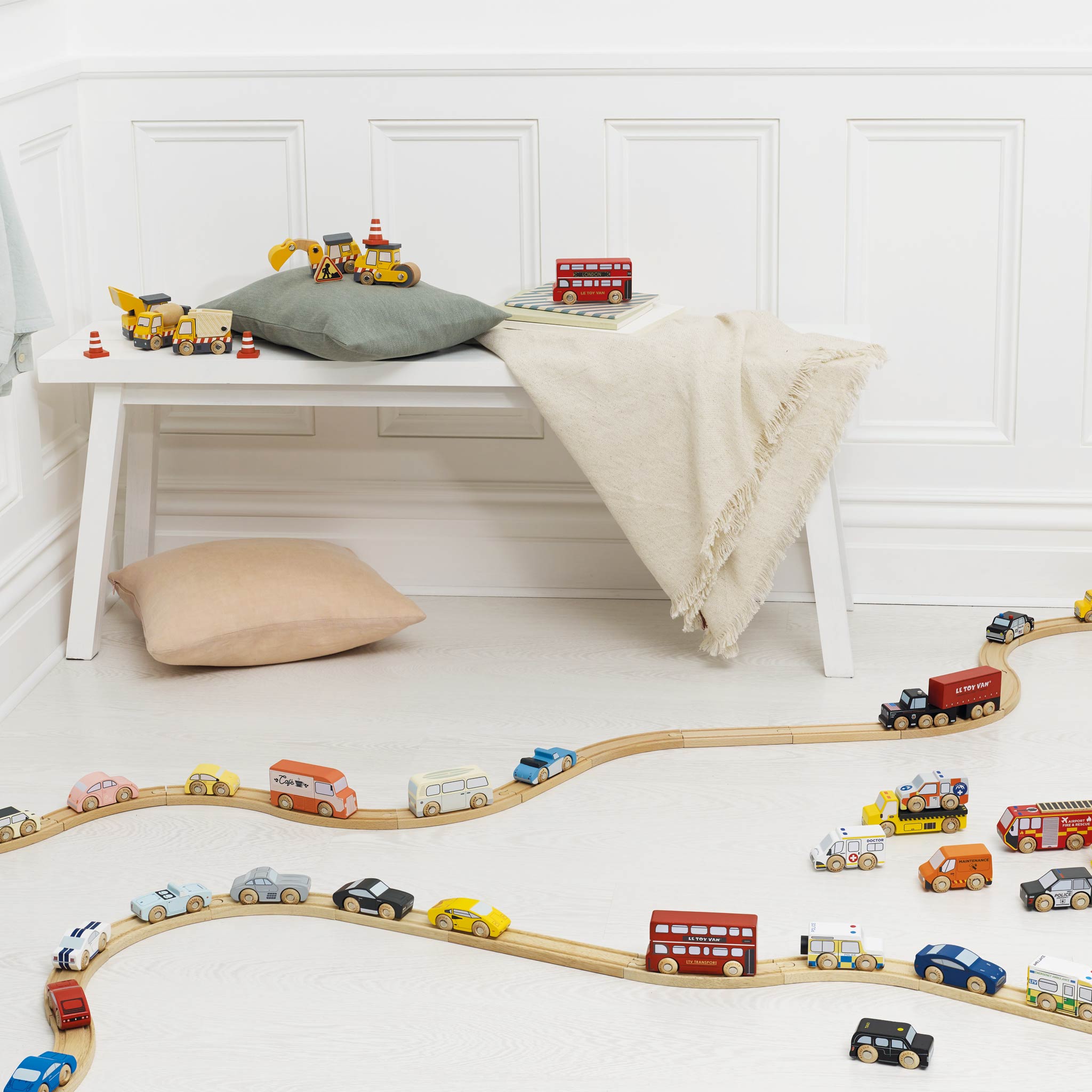 Wooden toy train set on a white floor with a bench and pillows in the background.