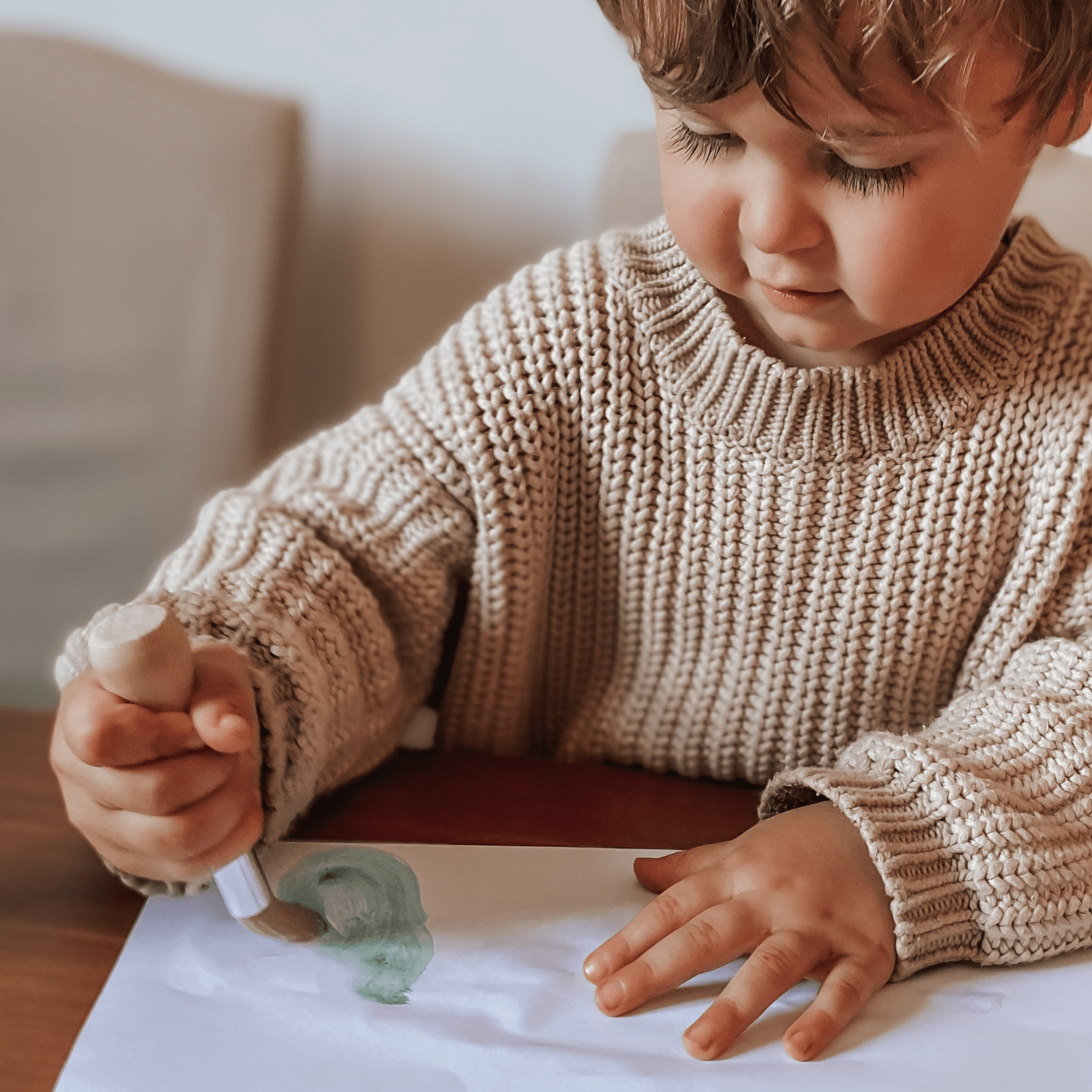 Child in a beige sweater playing with play dough on a table.