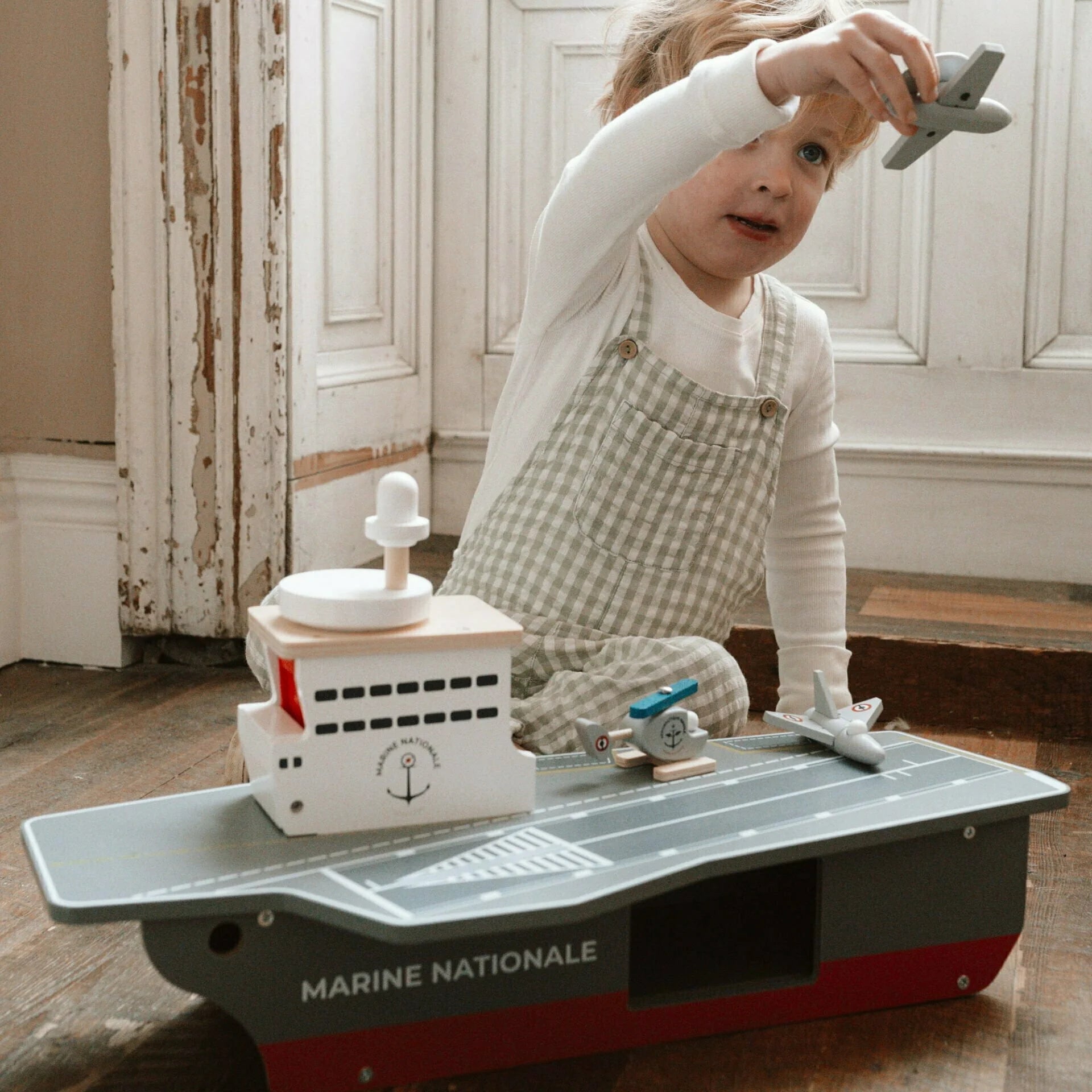 Child playing with toy airplane and ship on a large toy boat labeled 'Marine Nationale'.
