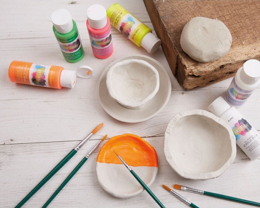 Ceramic bowls, paint bottles, and brushes on a wooden surface