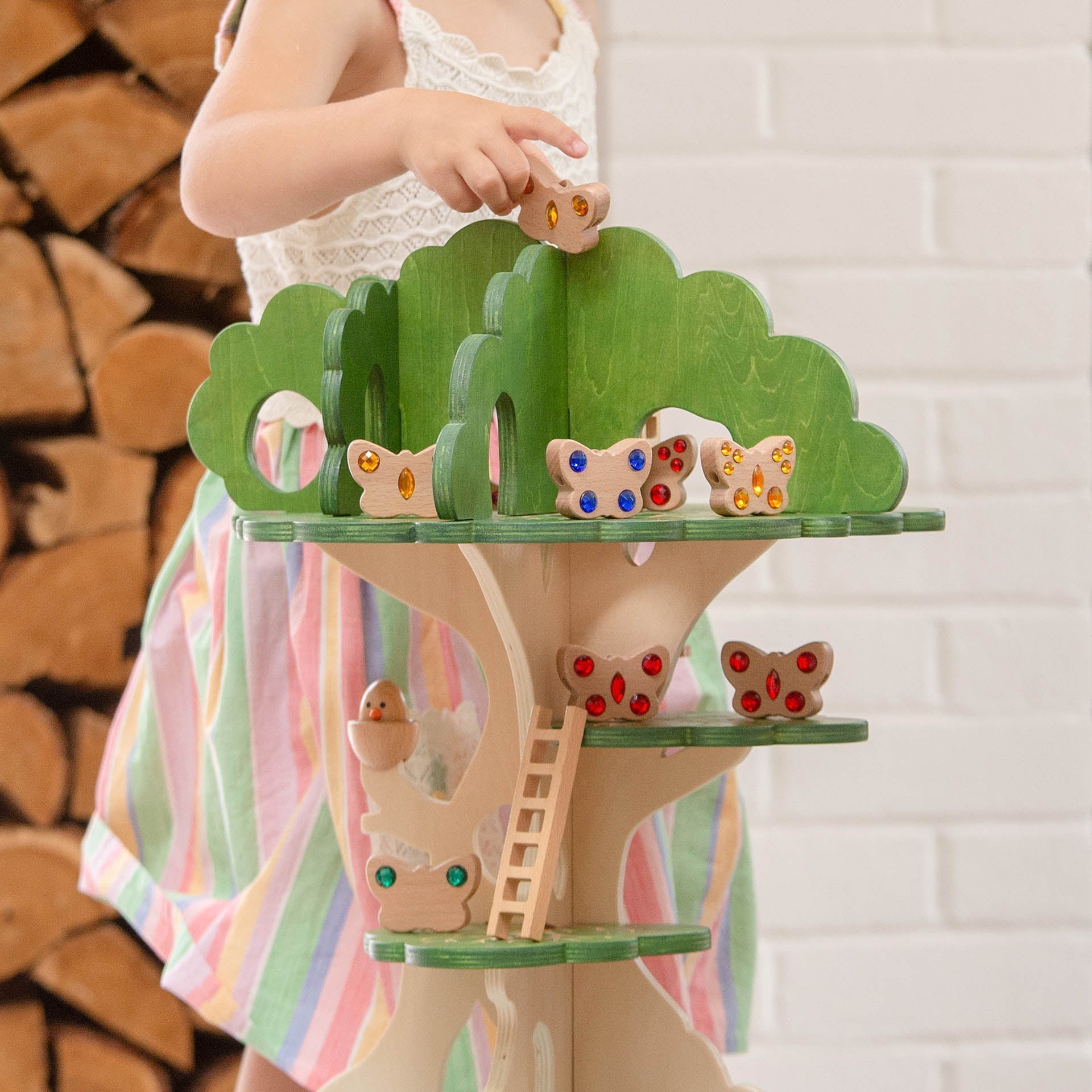 Child holding a colorful wooden treehouse toy with butterflies on a white brick wall background.