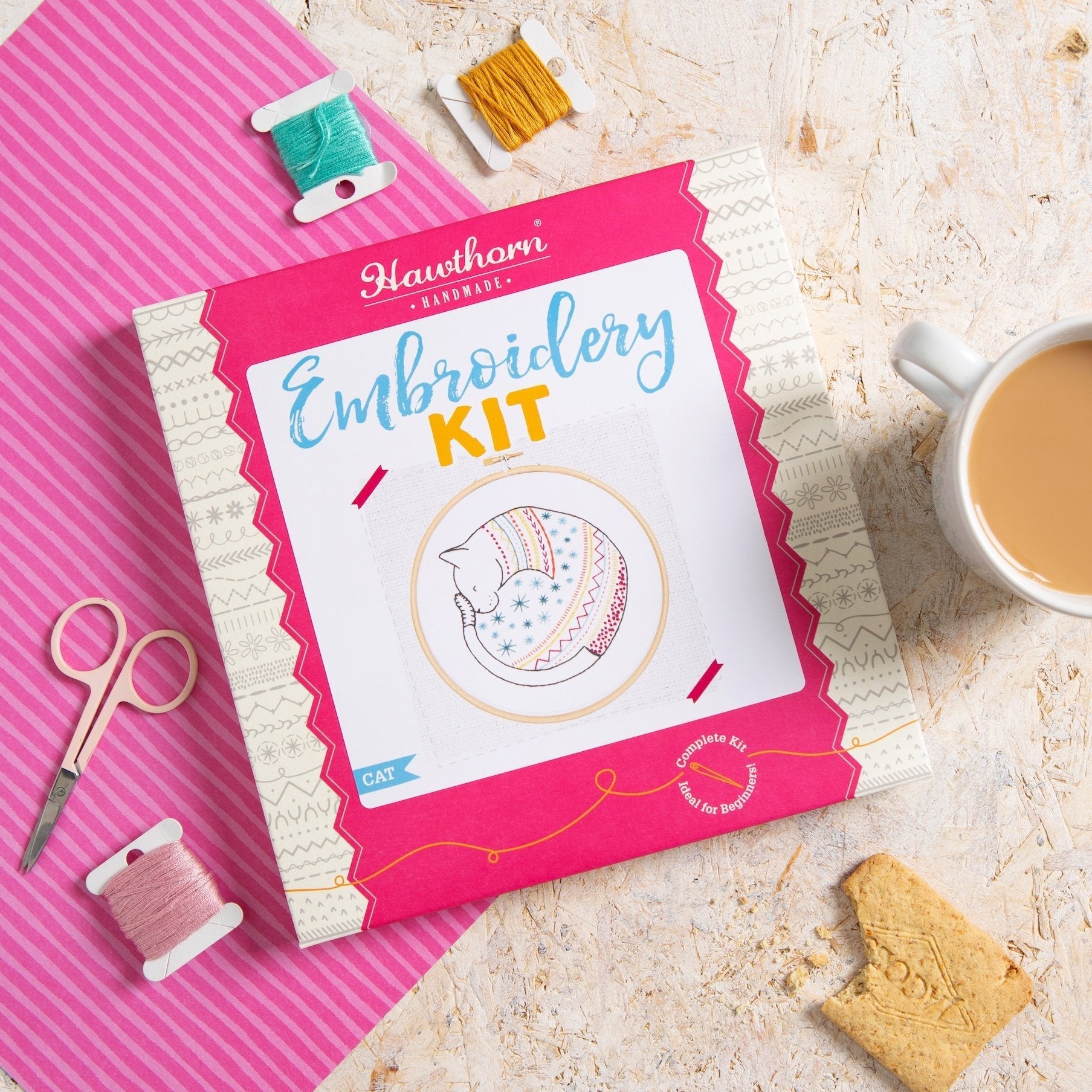Embroidery kit with thread spools, scissors, and a cup of tea on a pink and white striped surface.
