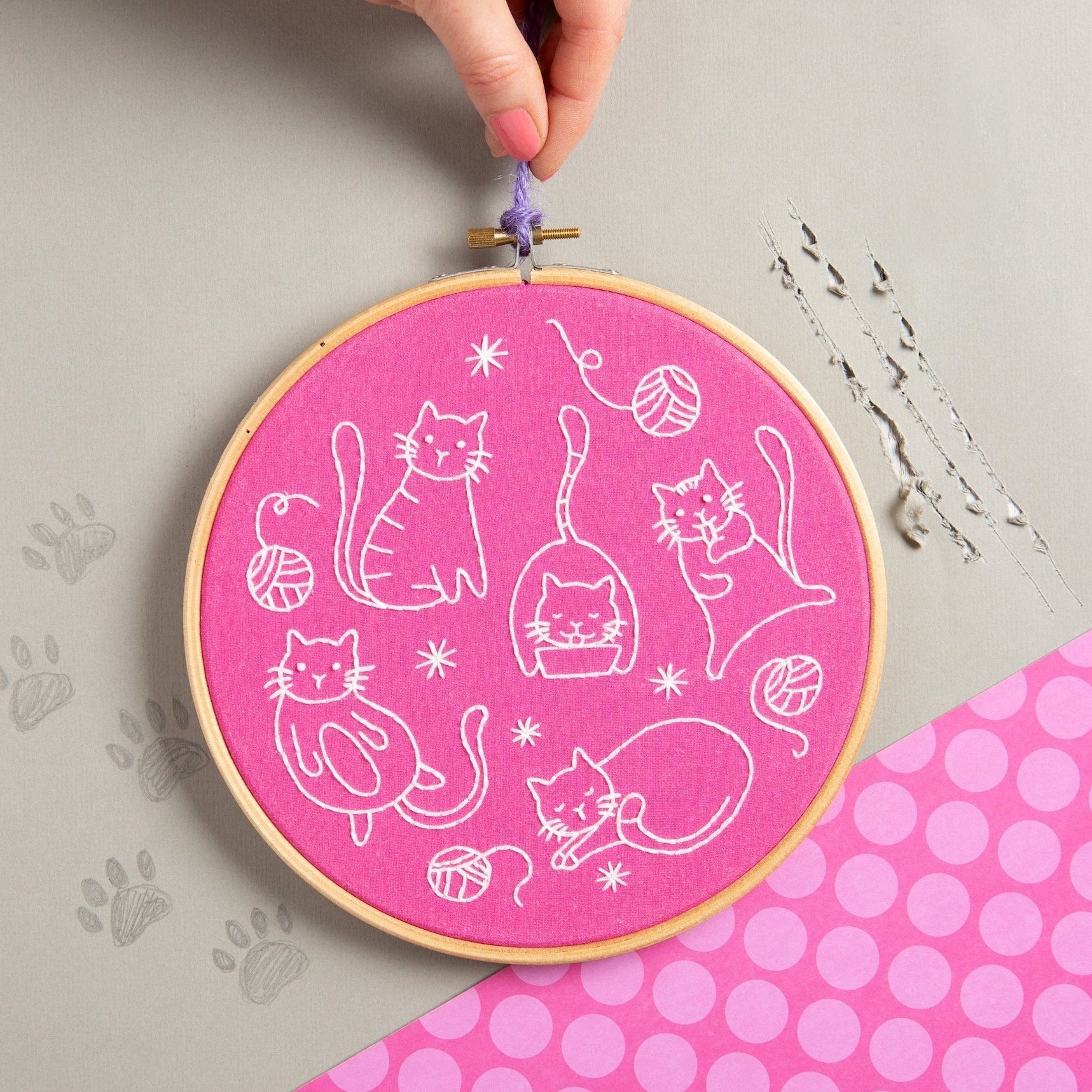 Embroidery hoop with white cat designs on a pink background, held by a hand.