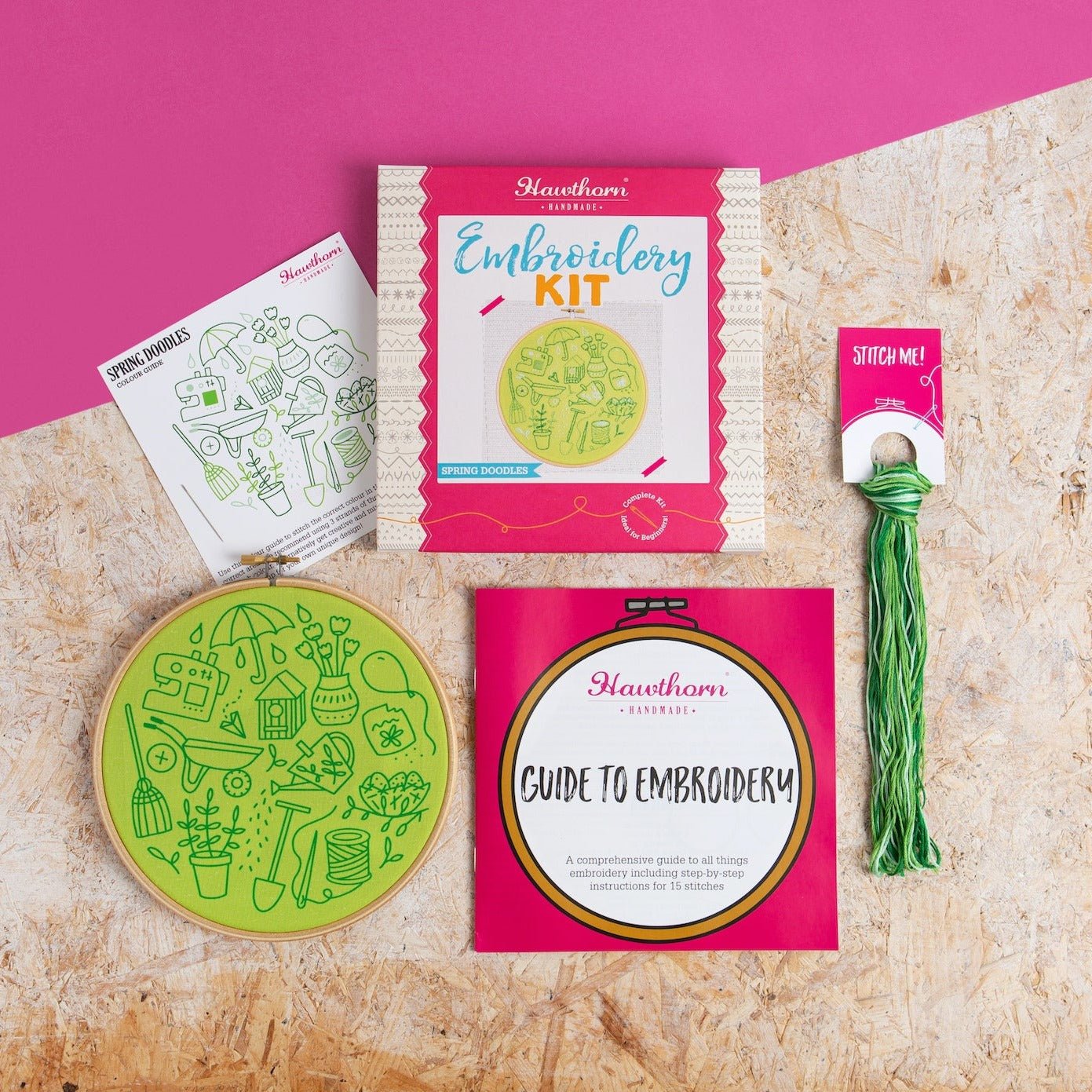 Embroidery kit with pink packaging, green embroidery hoop, and guide on a textured surface.