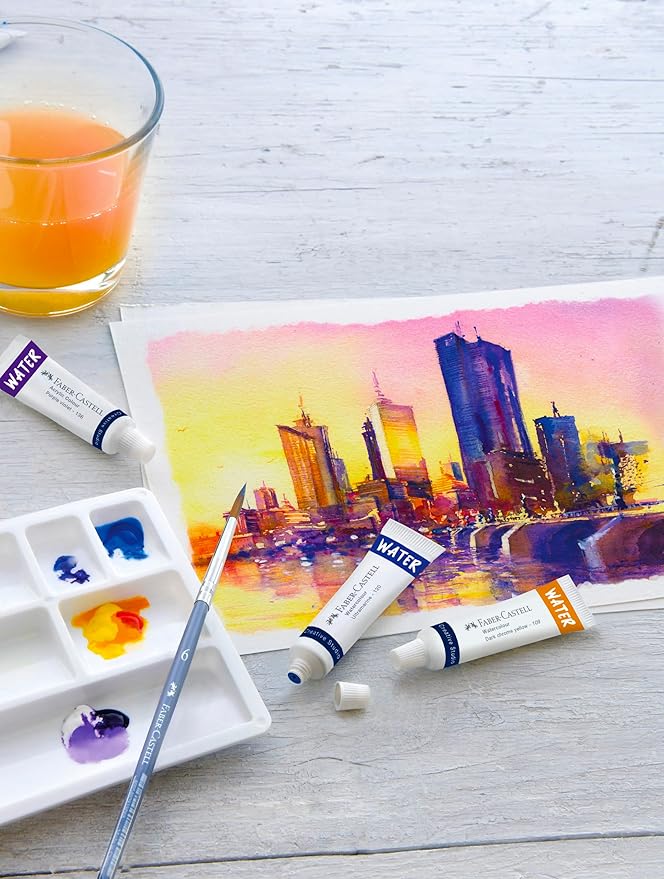 Artistic setup with watercolor painting of a cityscape, tubes of watercolor paint on a light wooden surface.