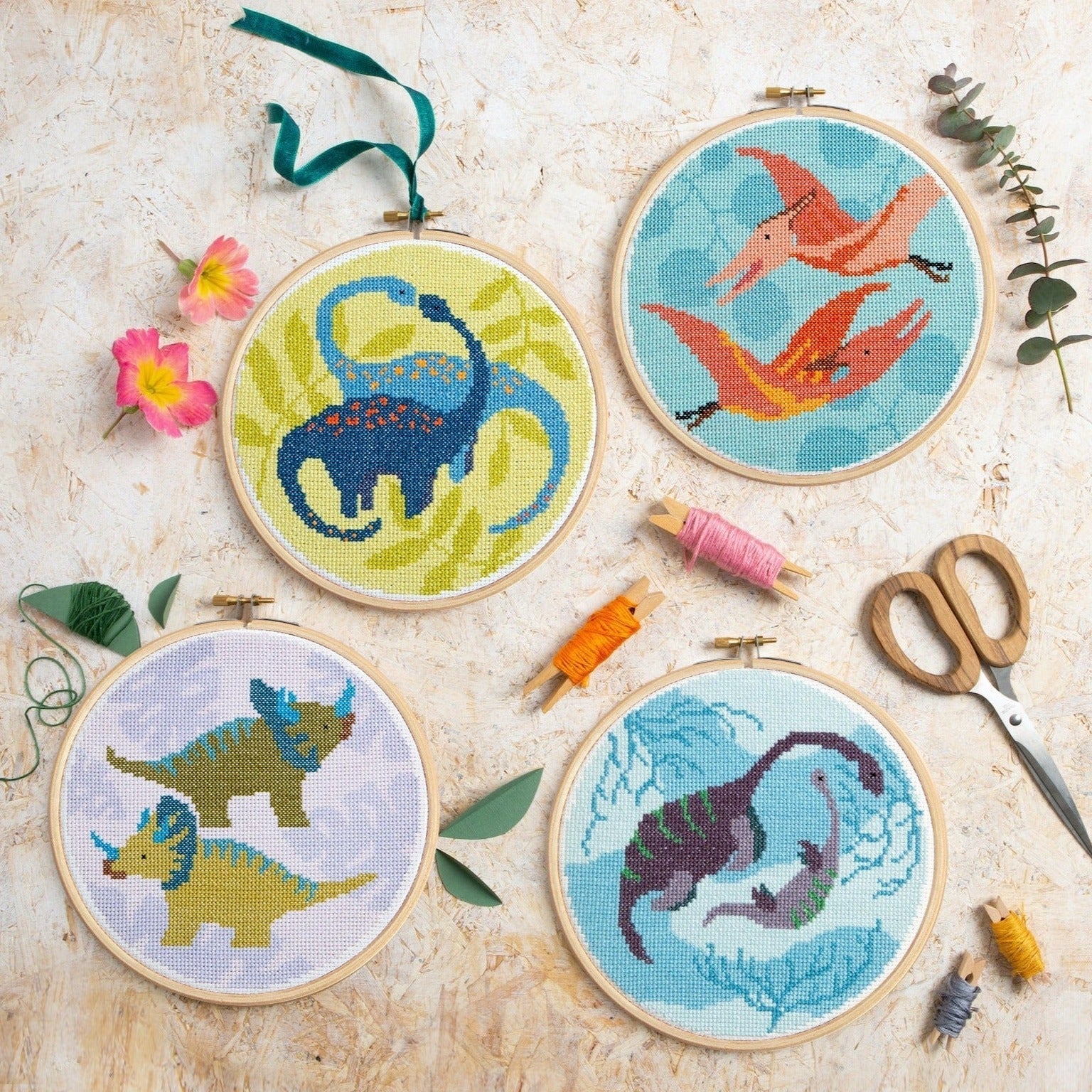 Four cross-stitched dinosaur designs in hoops on a textured surface with sewing tools.