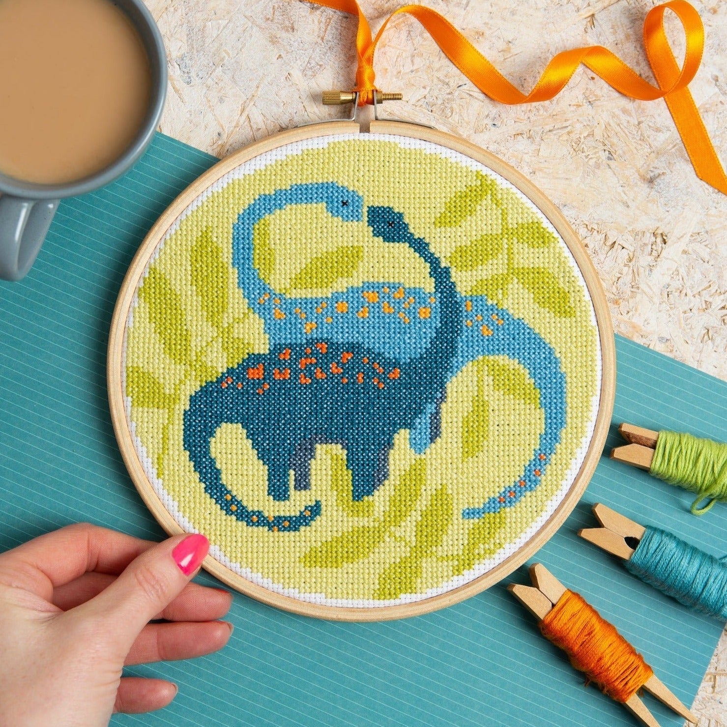 Embroidery hoop with a blue and green design held by a hand, surrounded by embroidery supplies on a textured surface.
