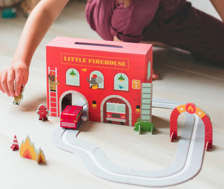 Children's toy set with a 'Little Firehouse' box, fire truck, and accessories on a light surface.