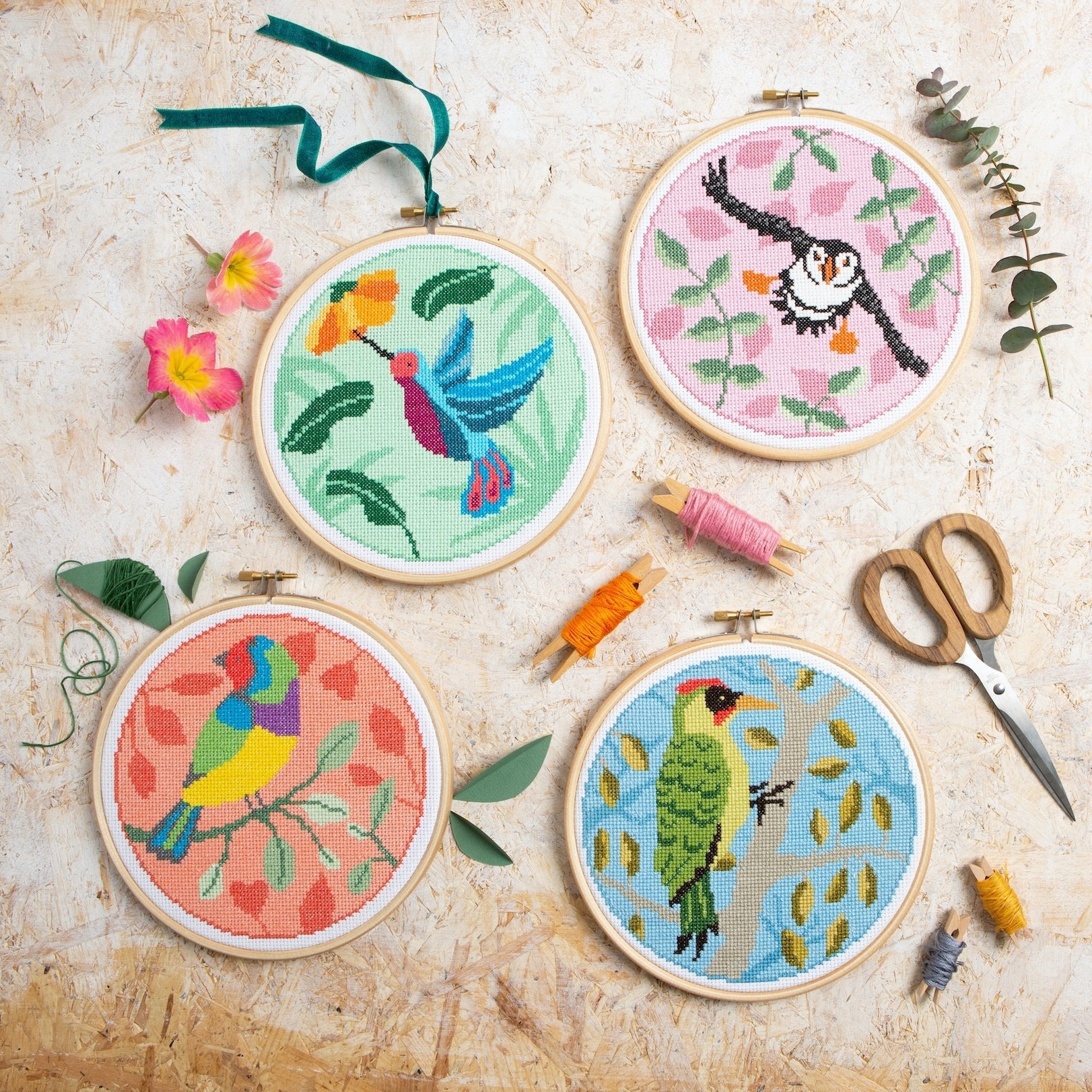 Four embroidered hoops with colorful bird designs on a textured surface with embroidery tools.