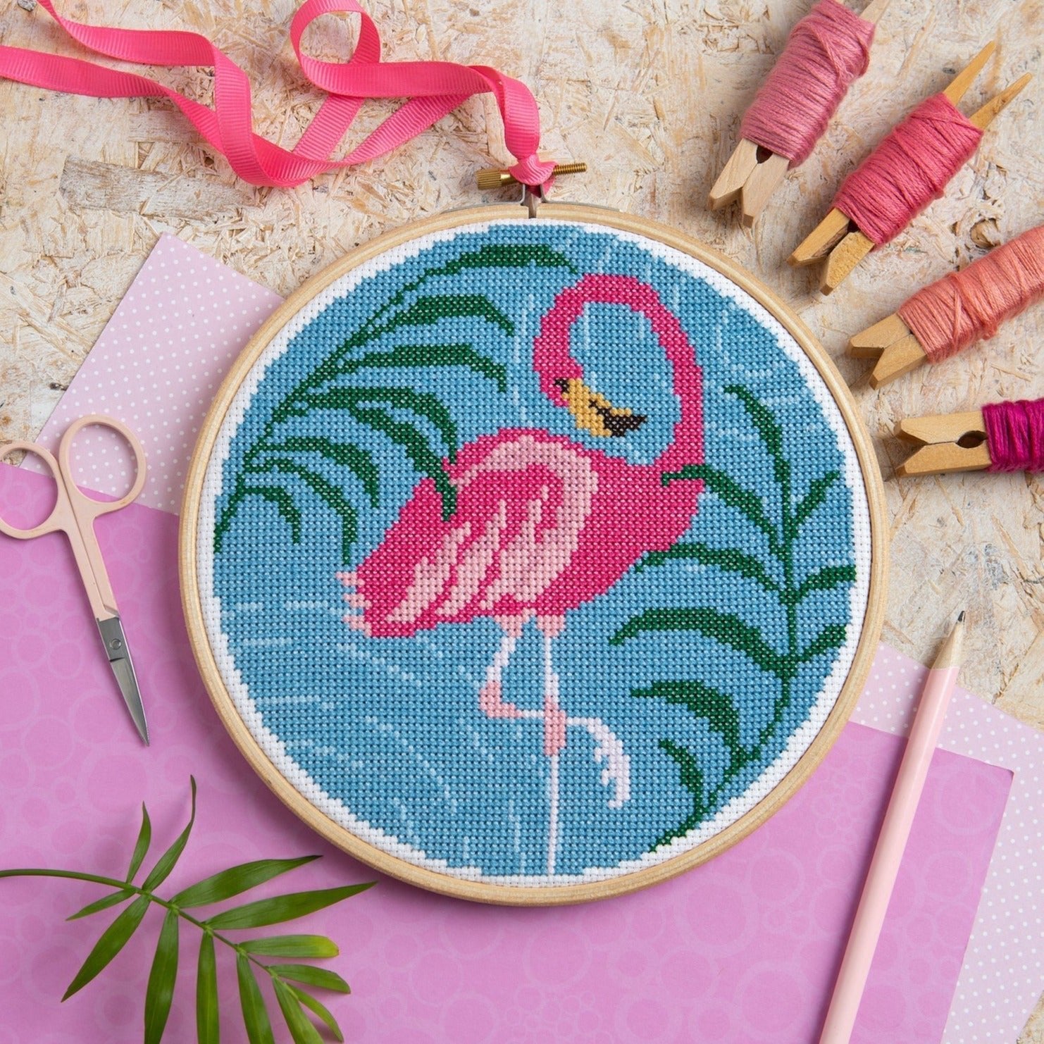 Cross-stitched flamingo design in a hoop with embroidery tools on a pink surface
