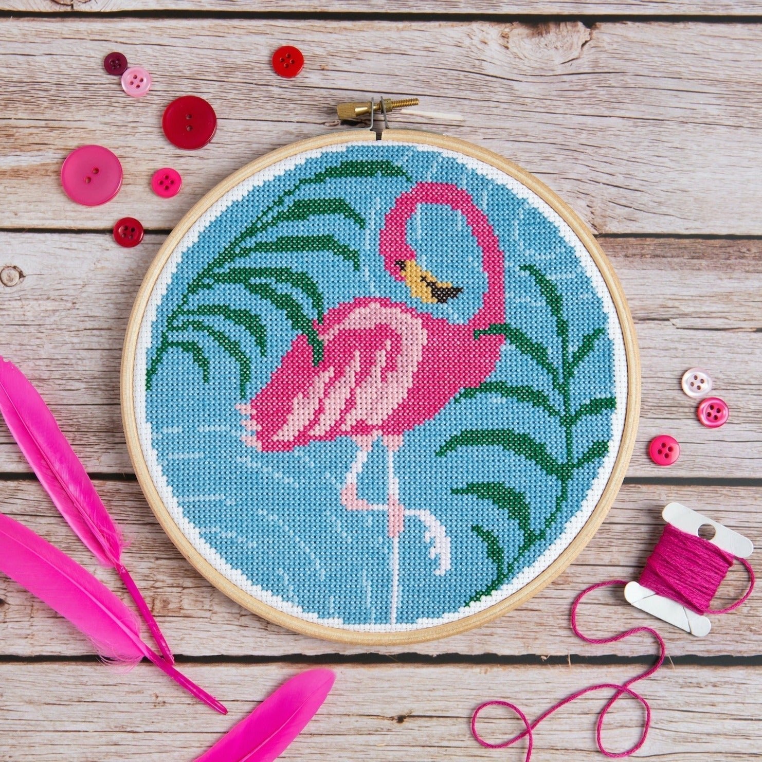 Embroidery hoop with a pink flamingo design on a wooden surface with buttons and feathers.
