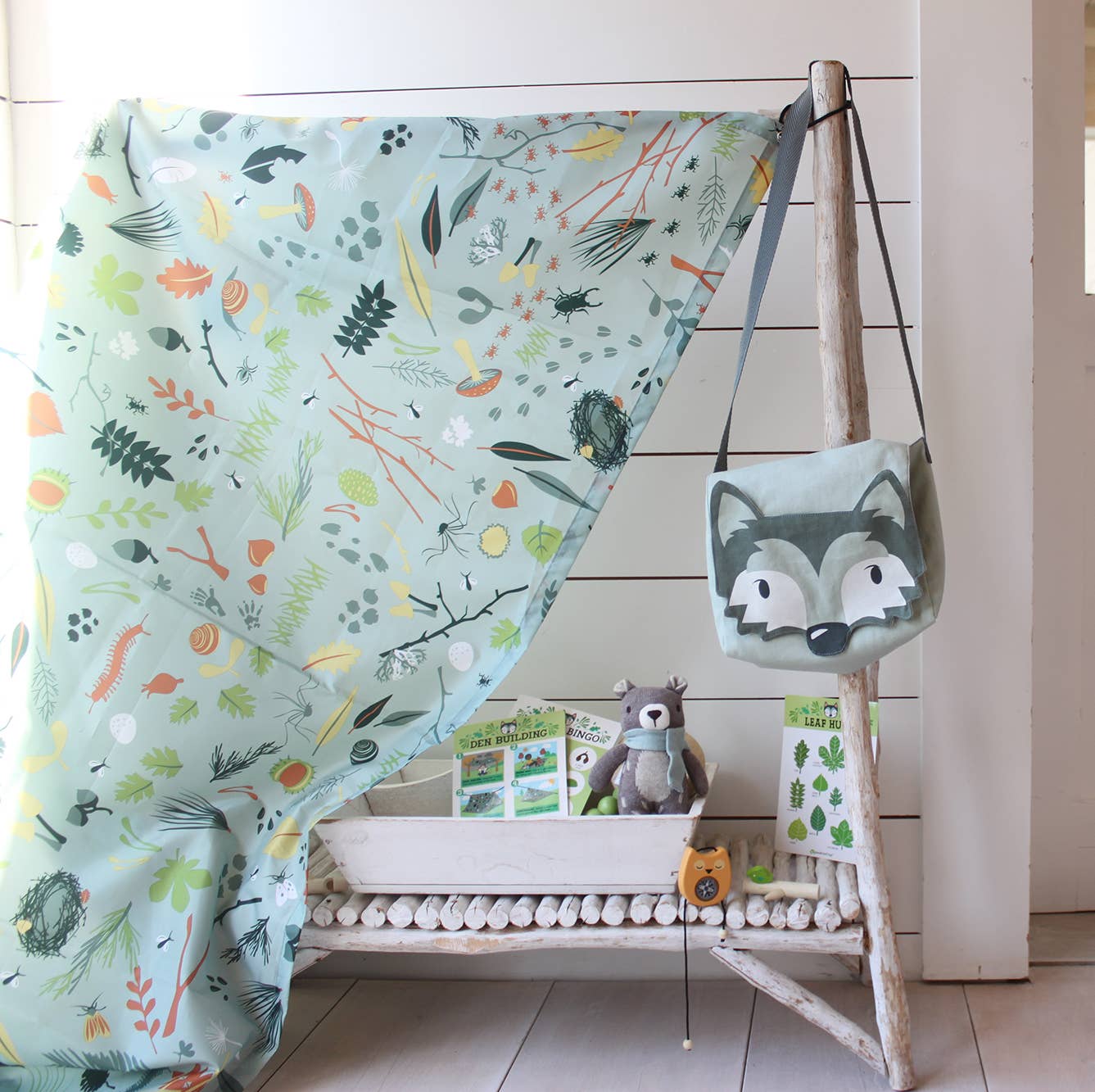 Children's room with a forest-themed blanket, toy bag, and books on a wooden stand.