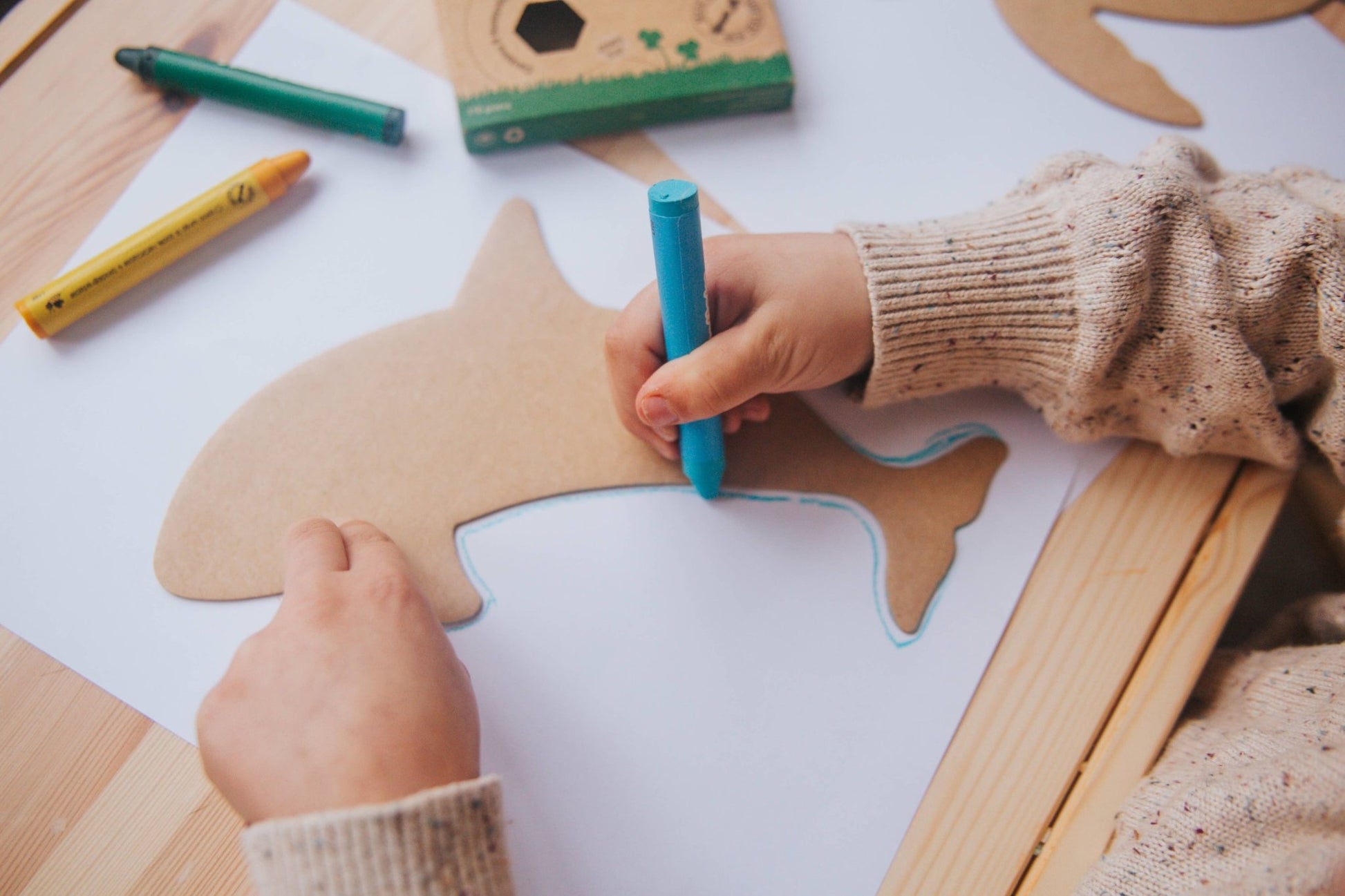 Child's hands coloring a shark-shaped paper cutout with a blue marker on a wooden table.