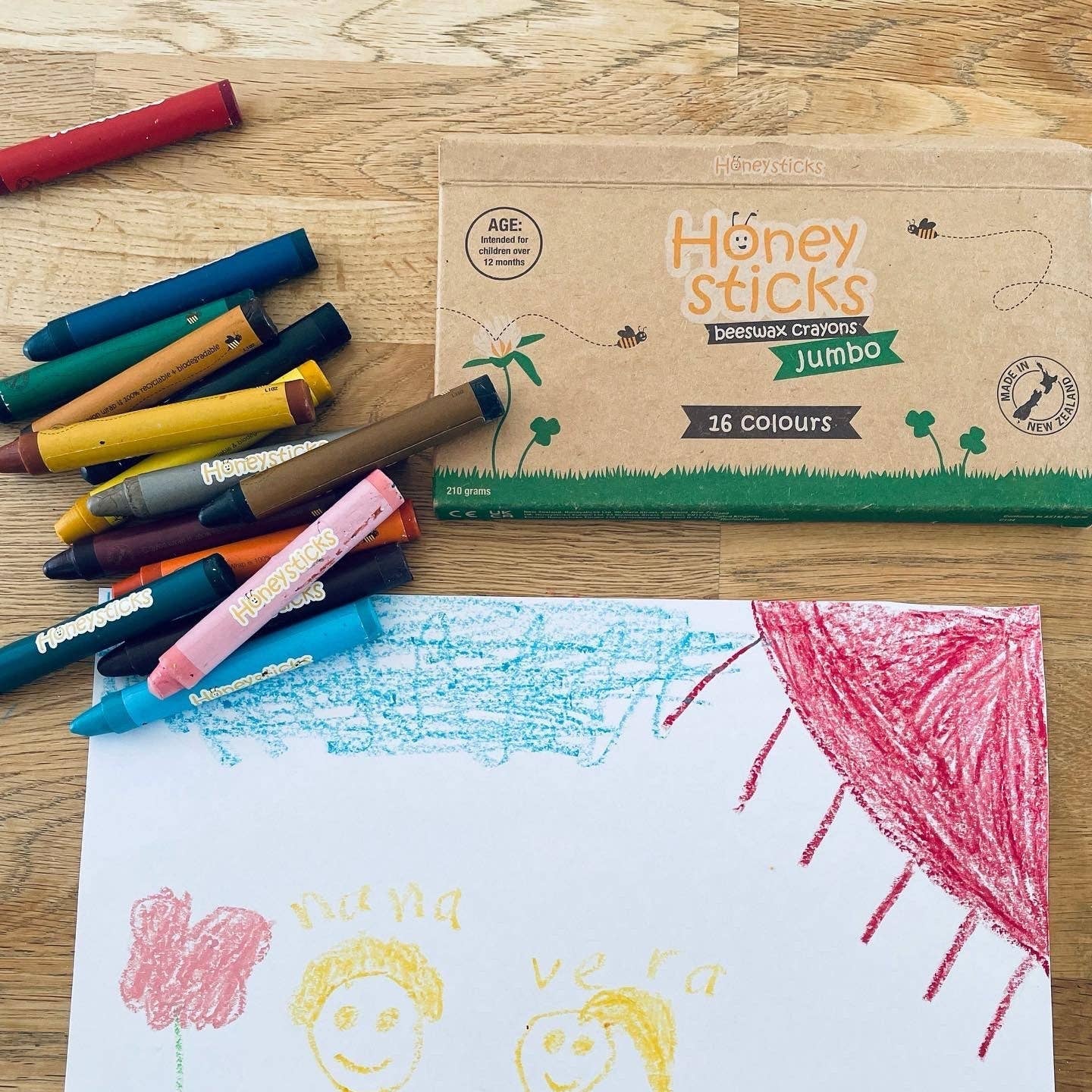 Children's drawing with crayons and a box of Honey Sticks crayons on a wooden surface.