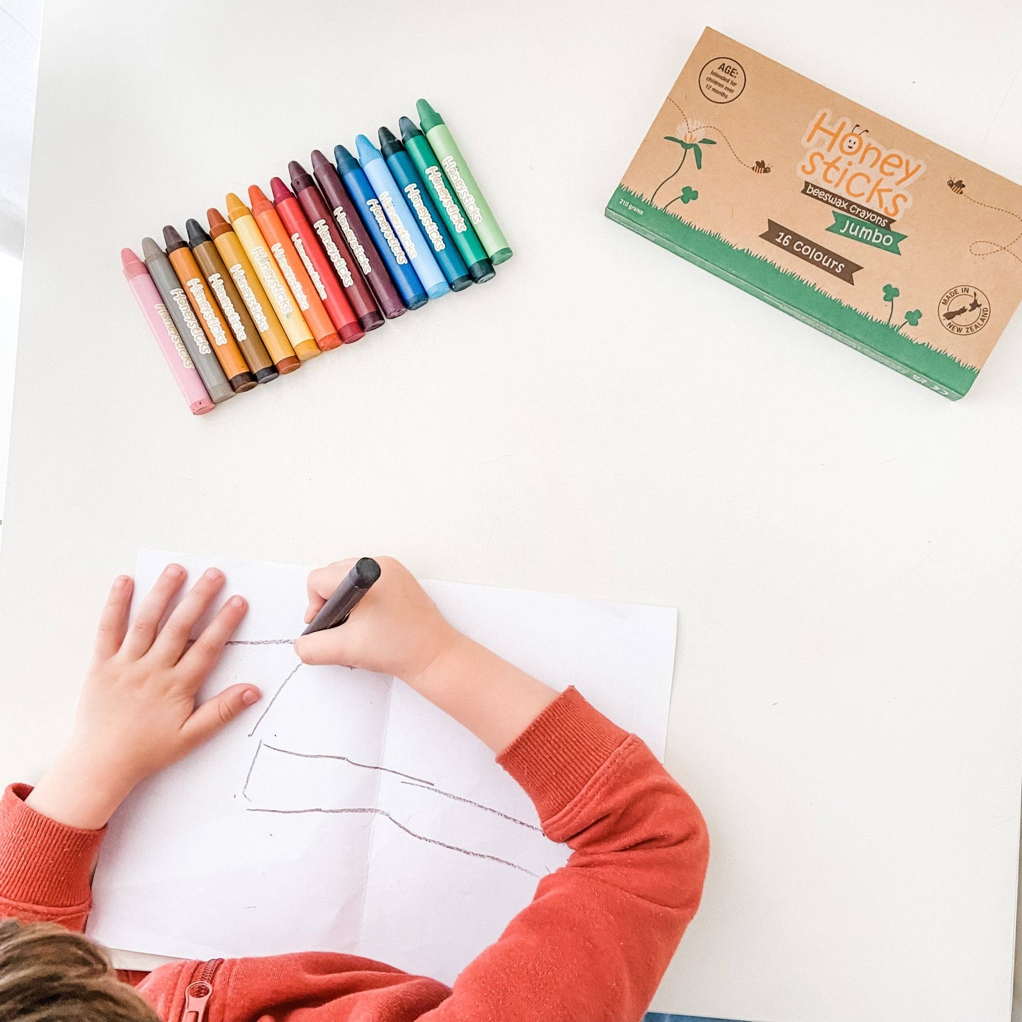 Child drawing with crayons on a white sheet of paper, with a box of crayons labeled 'Honey Sticks' in the background.