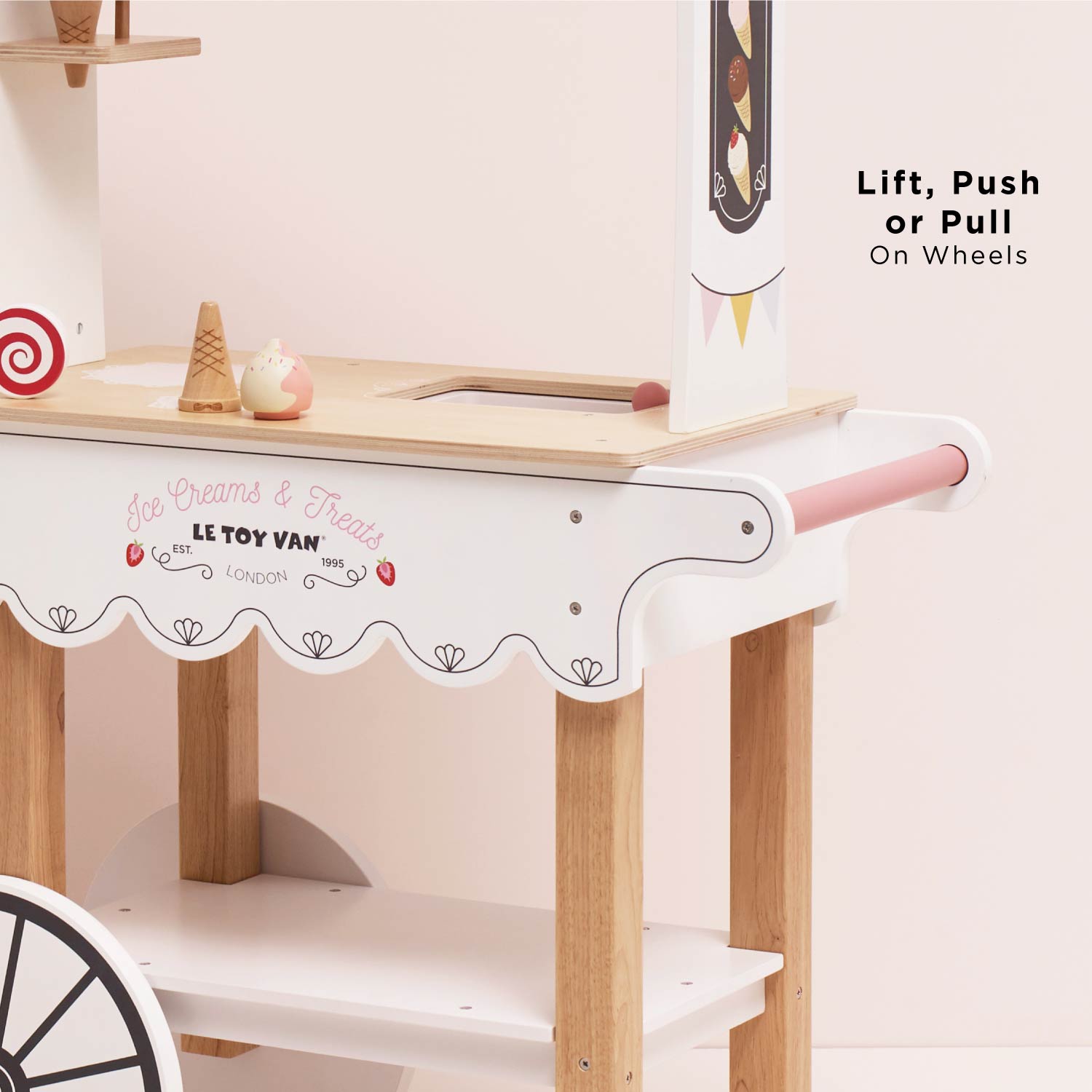 Wooden toy ice cream truck with wheels and a pink handle on a light pink background.