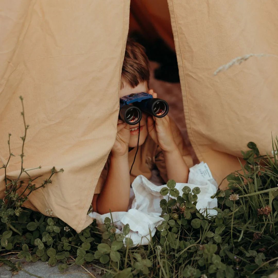 Person peeking out from a tent with binoculars, surrounded by grass and plants