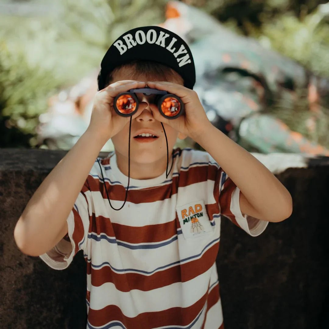 Child wearing a 'Brooklyn' cap and striped shirt, looking through binoculars with a blurred natural background.