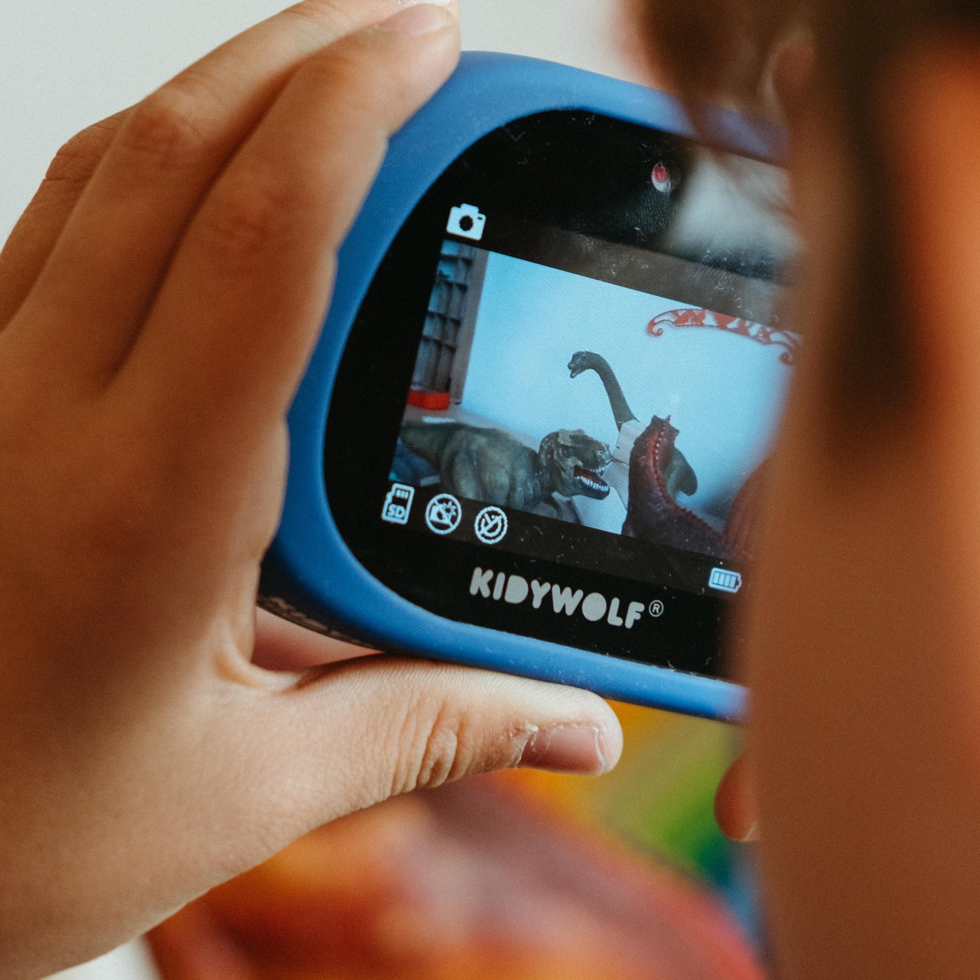Child holding a blue toy camera with a screen displaying cartoon characters, brand 'KIDYWOLF' visible.