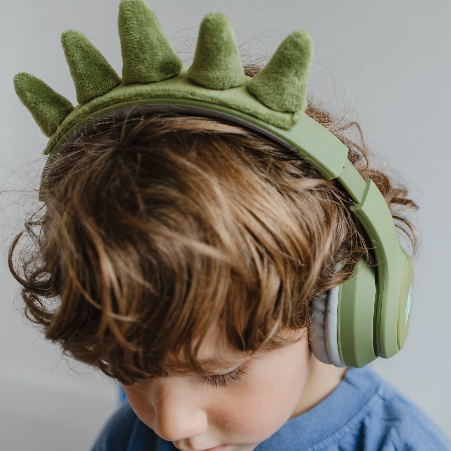Child wearing green dinosaur-shaped headphones with a plain background