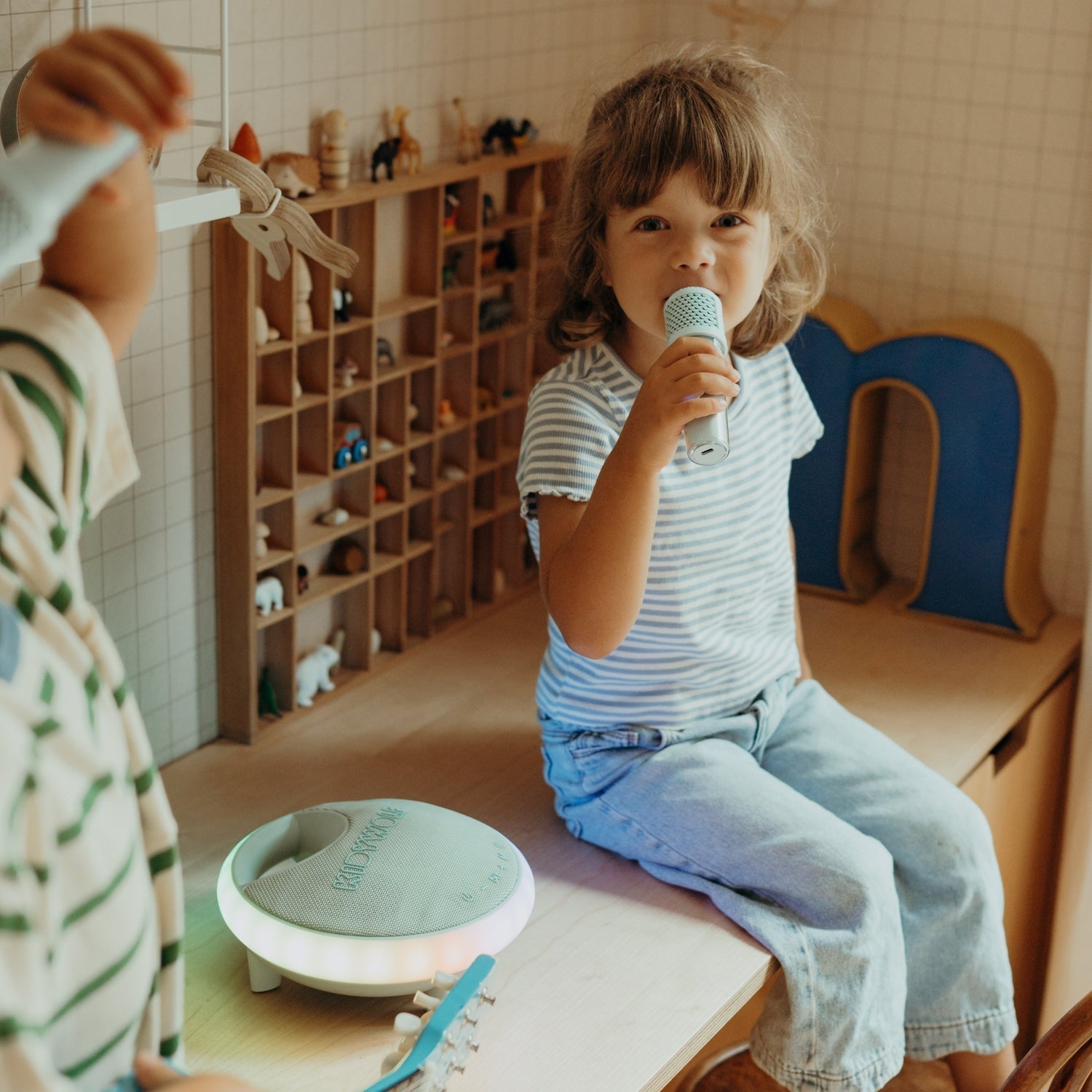 Child holding a microphone in a classroom setting with educational toys and furniture.