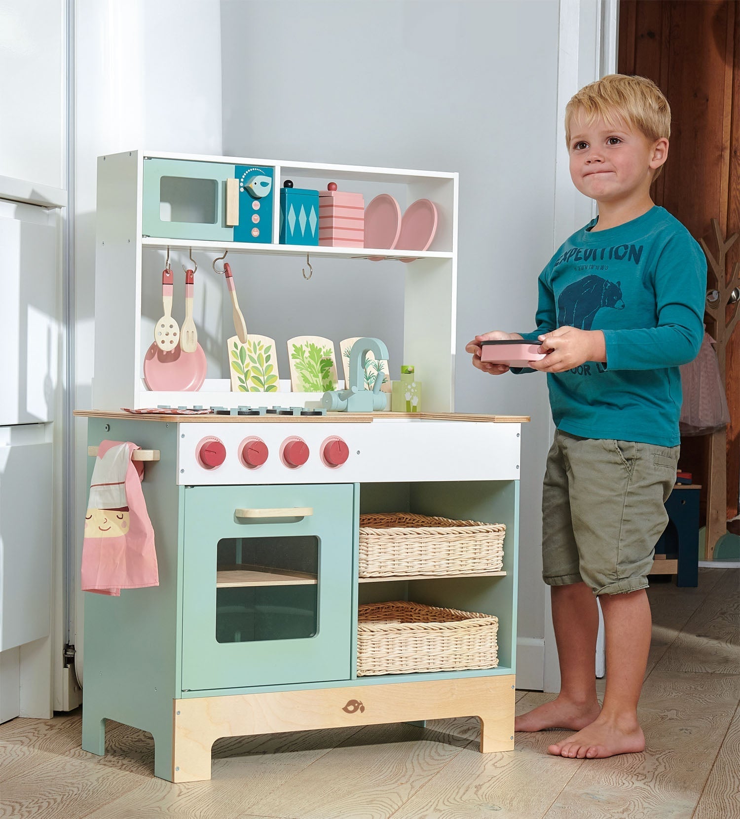 Child standing next to a toy kitchen set in a room.