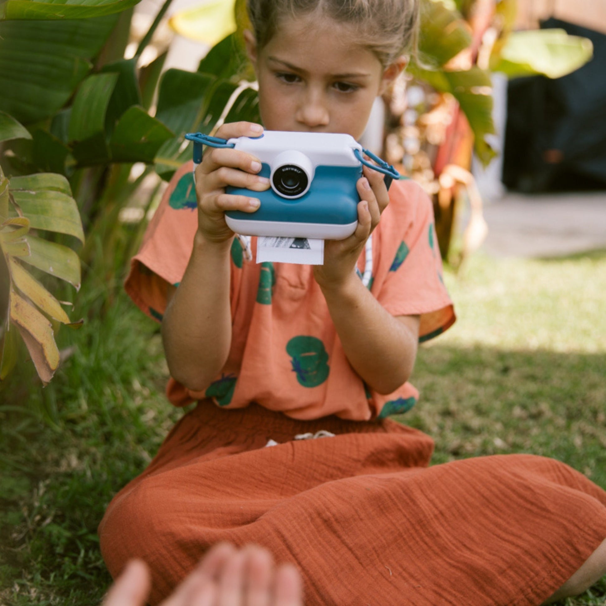 Child holding a blue and white camera outdoors with greenery in the background