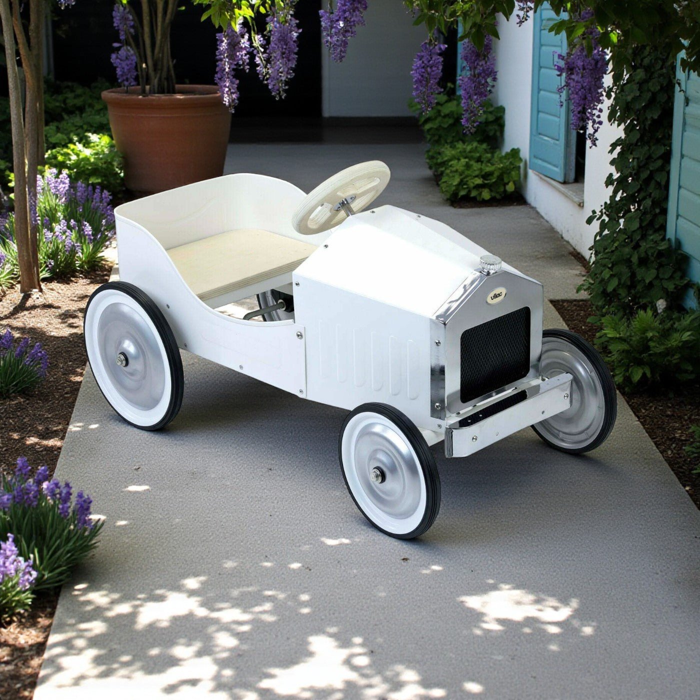 White vintage-style child's toy car on a concrete path with plants and flowers in the background.
