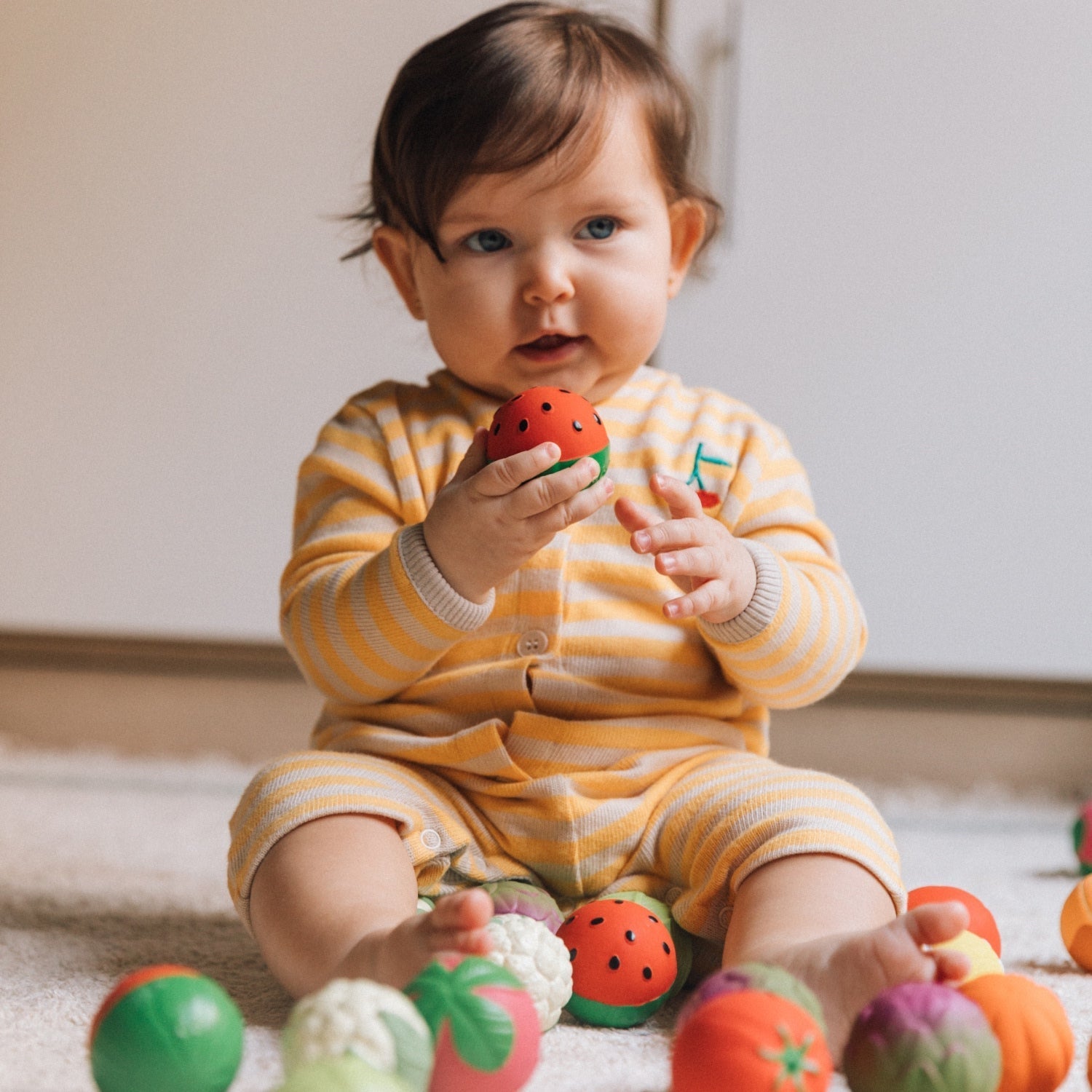 Baby in yellow striped outfit holding a red toy, surrounded by colorful toys on a light wooden floor.