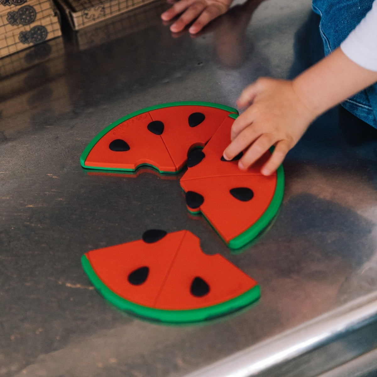 Children's watermelon puzzle on a metallic surface with a child's hand reaching for a piece.