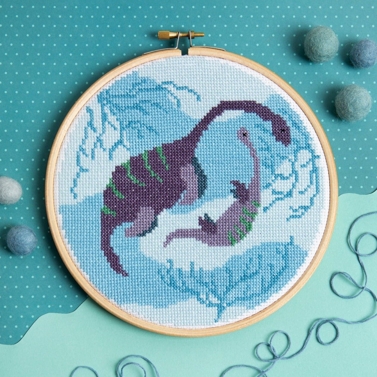 Embroidery hoop with a completed cross-stitch design of a purple dinosaur on a blue background.