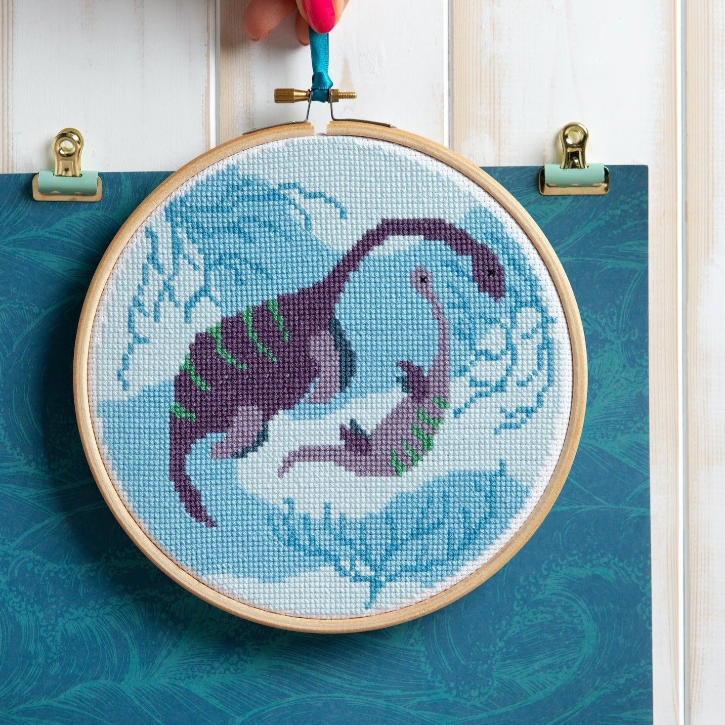 Embroidery hoop with a cross-stitched design of a purple dragon on a blue background.