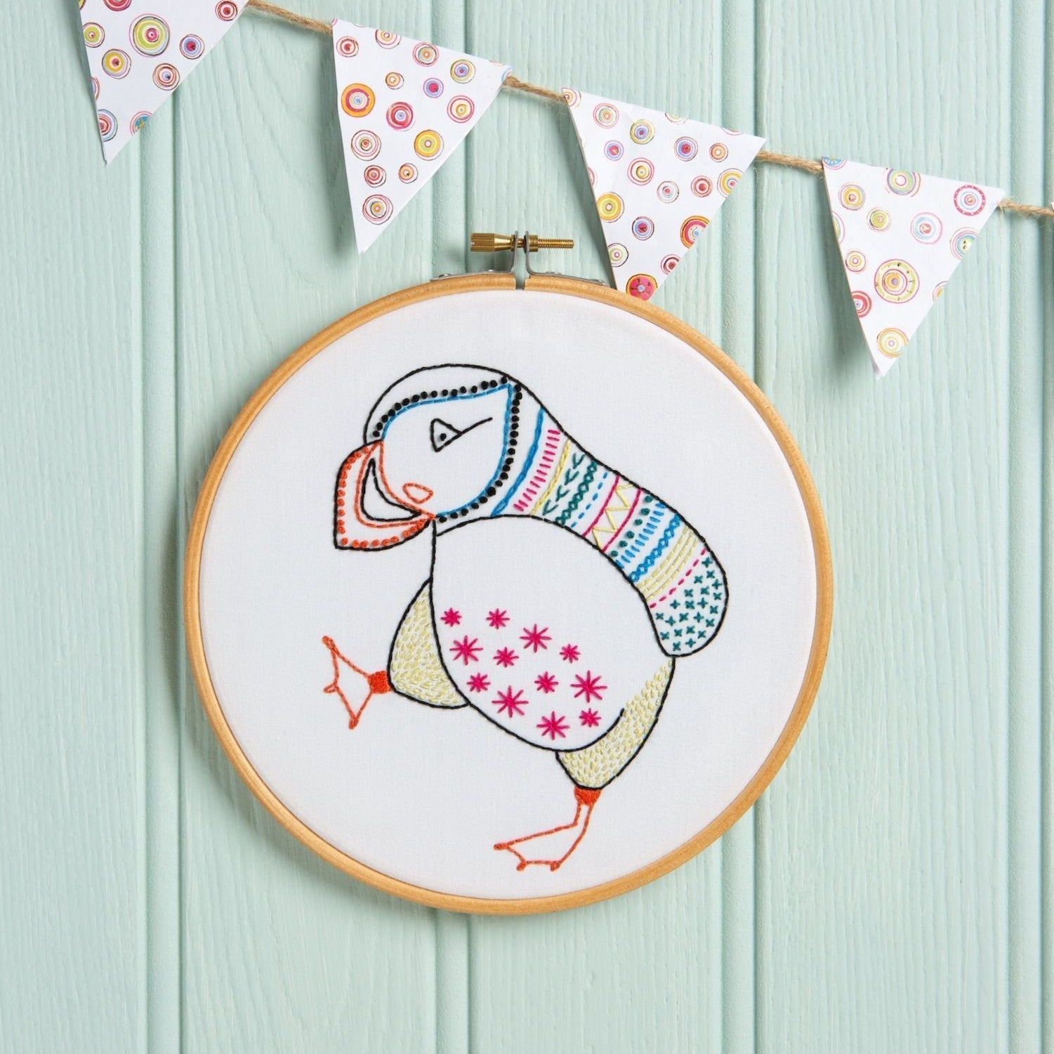 Embroidery hoop with a colorful puffin design on a light blue wooden background with decorative flags.