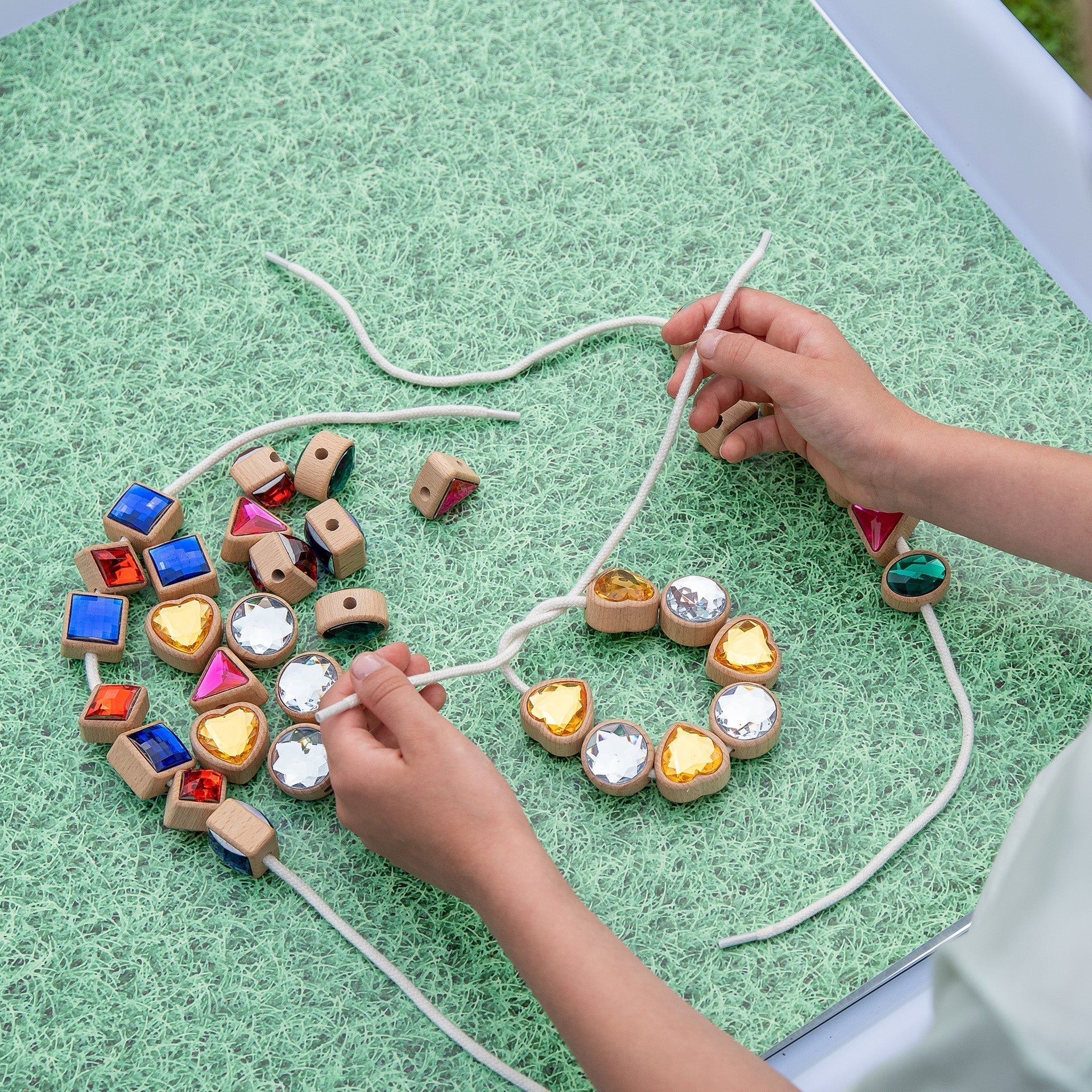 Children's hands threading colorful wooden beads on a string over a green surface.