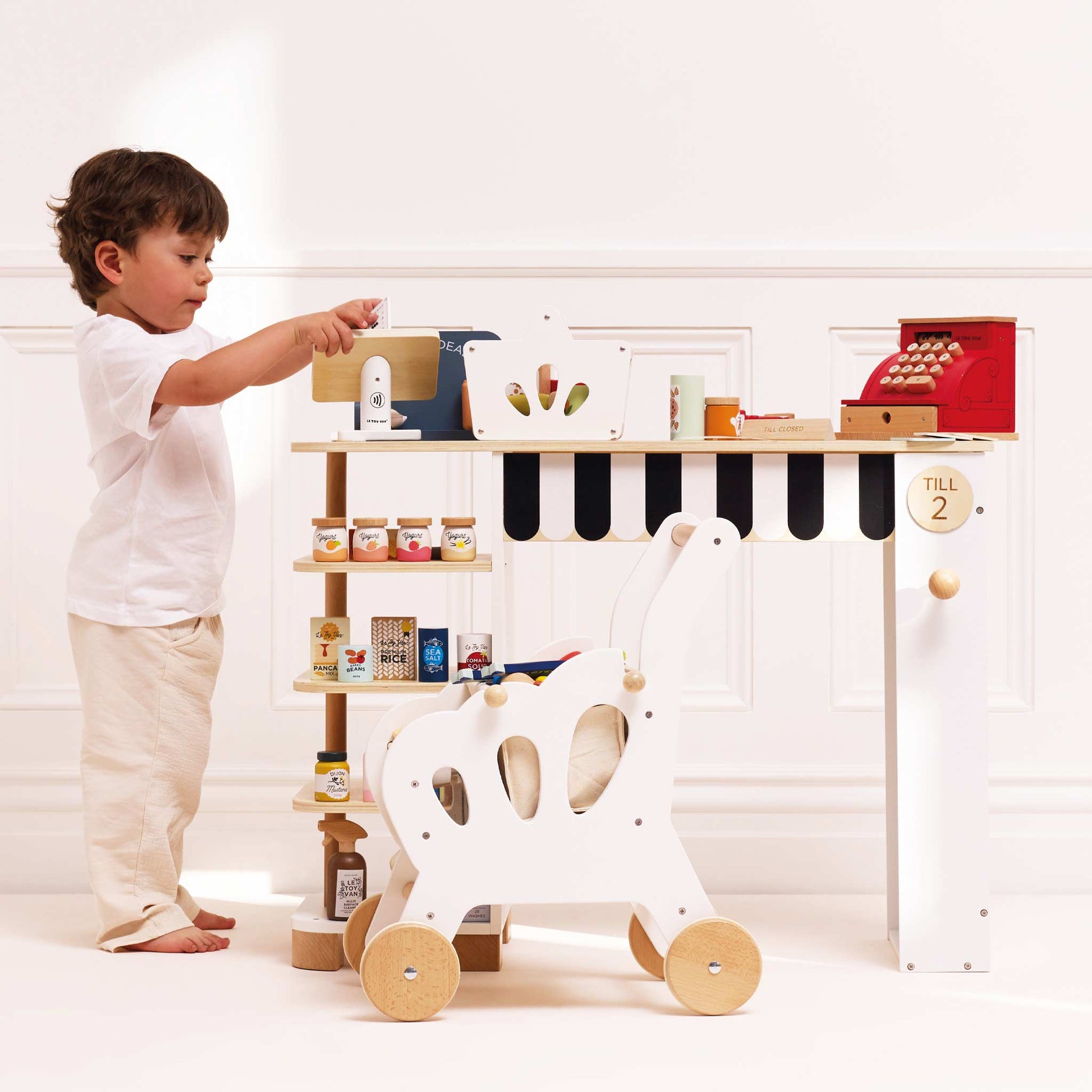 Child playing with a toy cash register and other items on a shelf in a bright room.