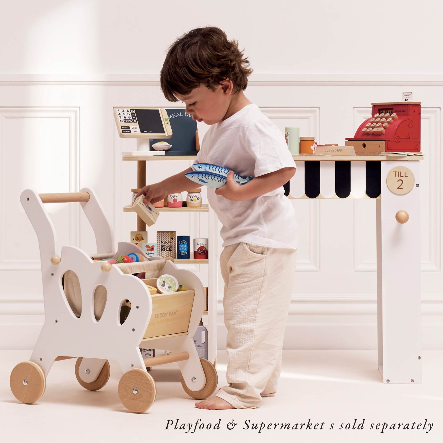 Child playing with a toy cash register and shopping cart in a white room.