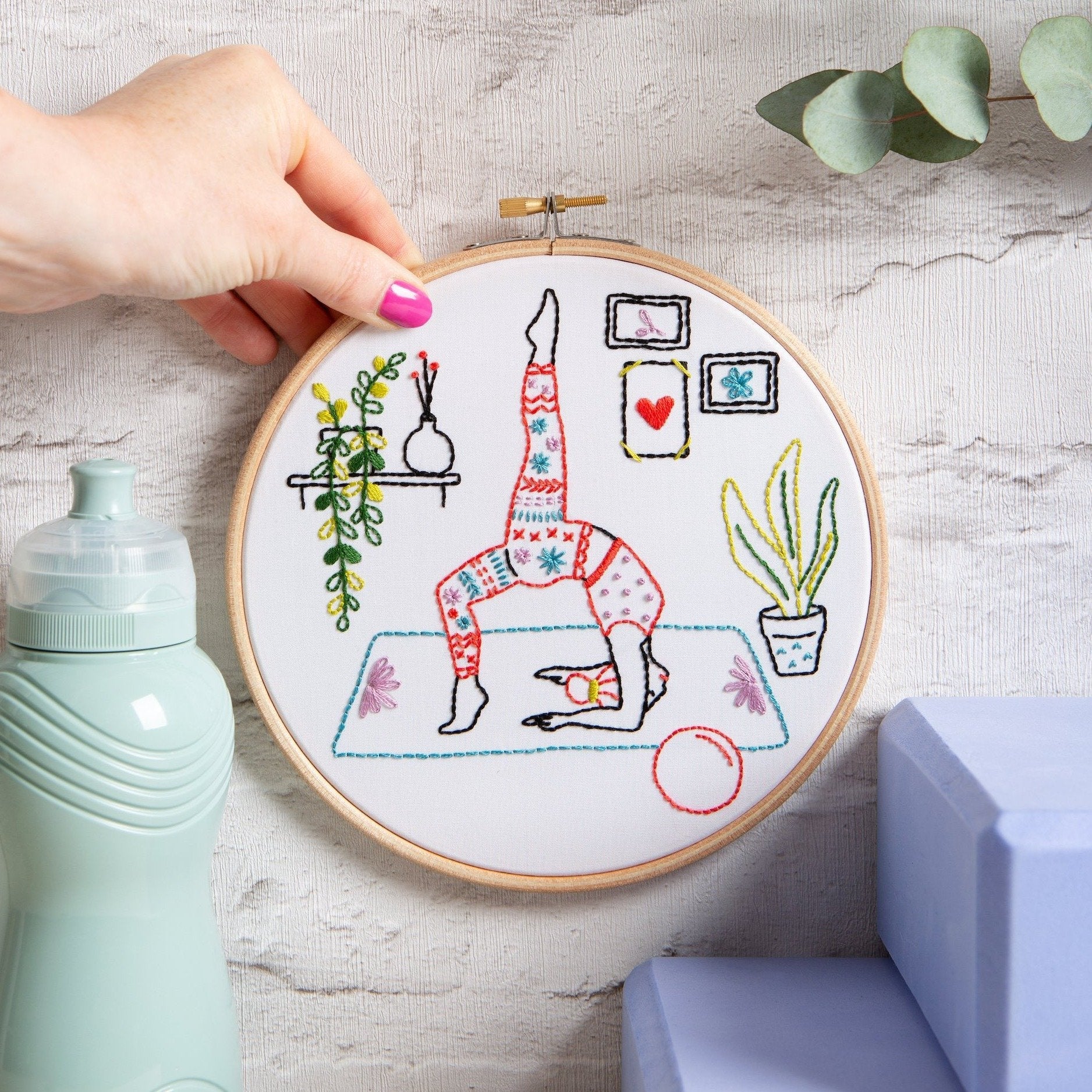Embroidery hoop with a colorful yoga design held by a hand on a light gray background.