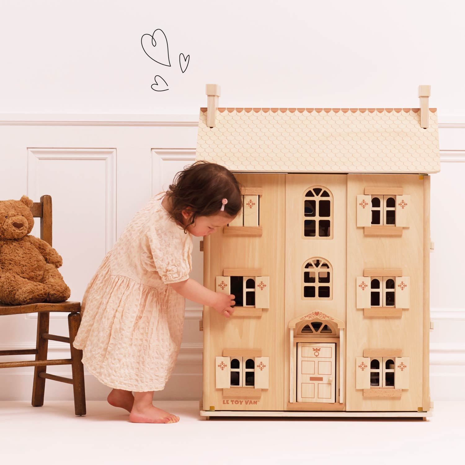 Child playing with a wooden dollhouse in a room with a teddy bear on a chair.