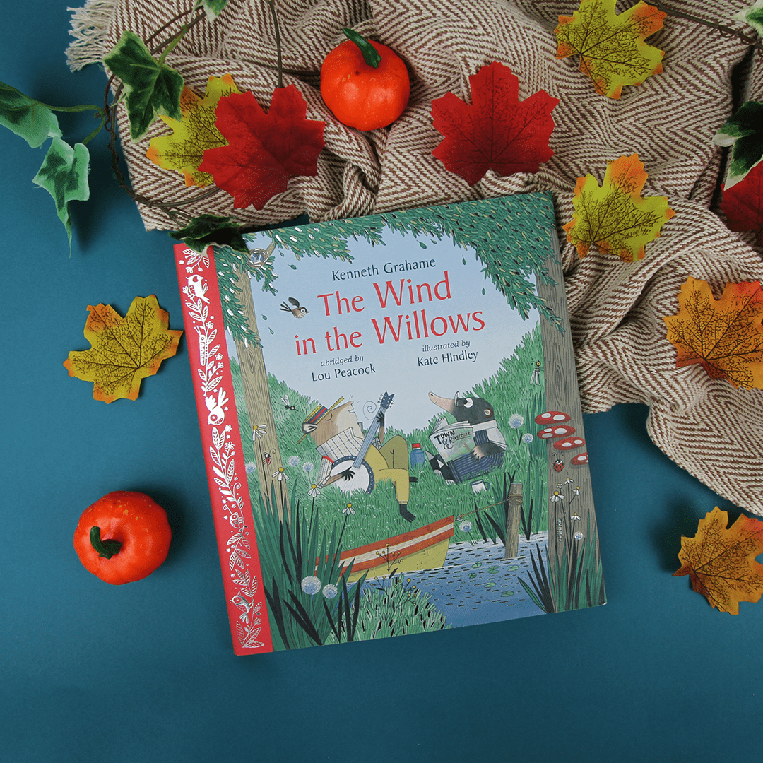 Book titled 'The Wind in the Willows' surrounded by autumn leaves and pumpkins on a textured surface.