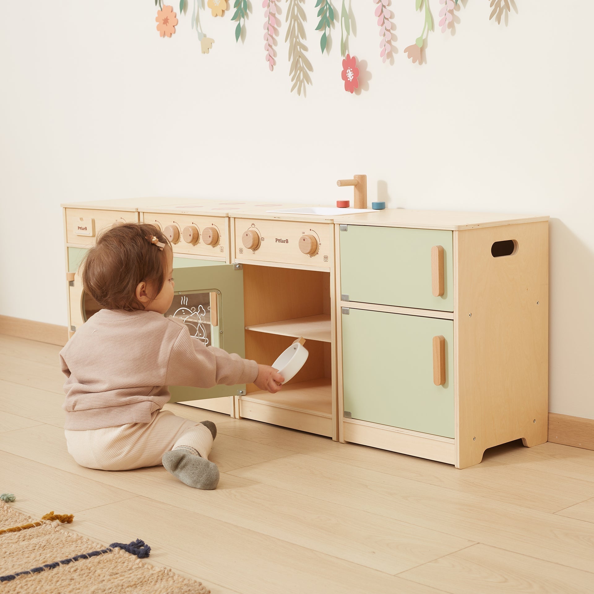Child playing with a wooden toy kitchen set in a room with floral decorations on the wall.