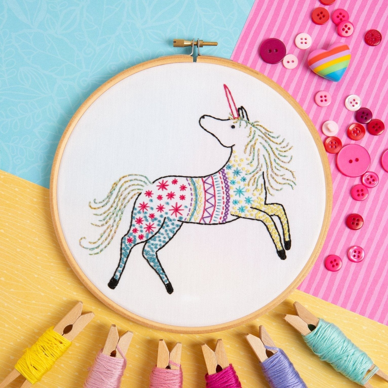 Embroidery hoop with a colorful unicorn design surrounded by sewing materials on a multicolored background