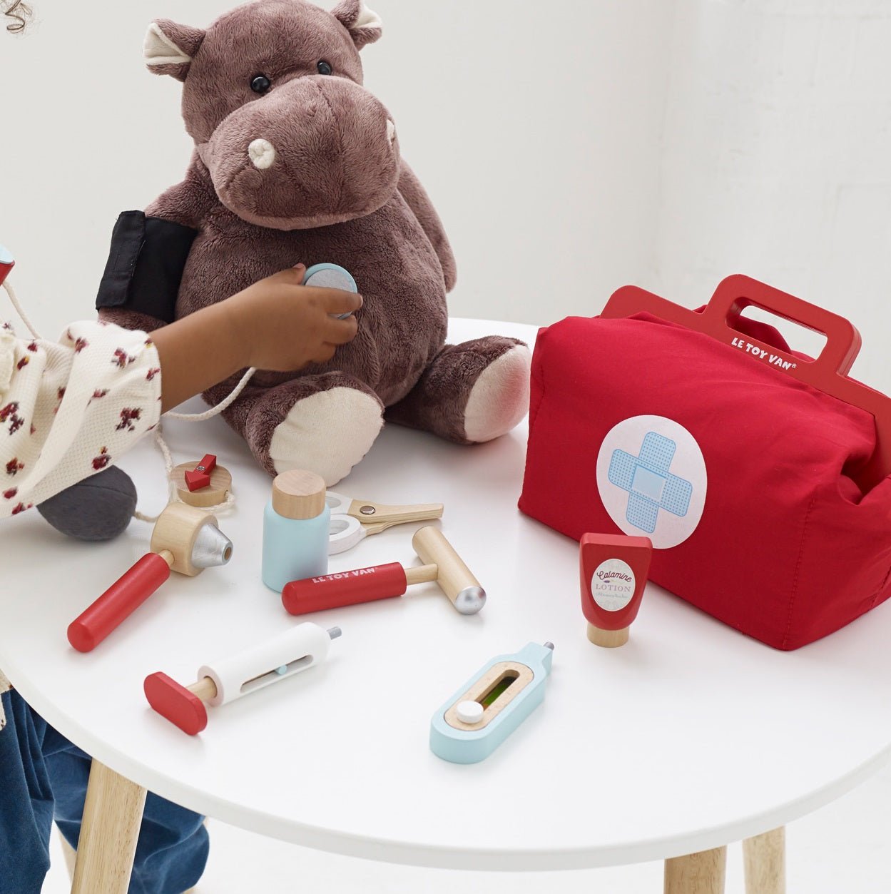 Children's toy doctor set with plush hippo, medical tools, and red bag on a white table.