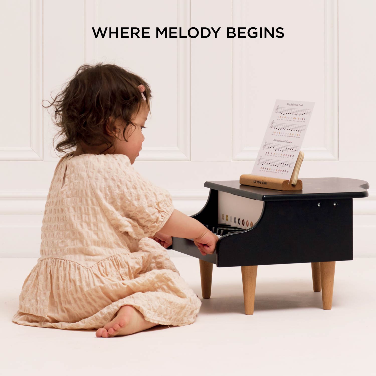 Child playing with a small piano-like toy with sheet music on a white background, featuring text 'WHERE MELODY BEGINS'.