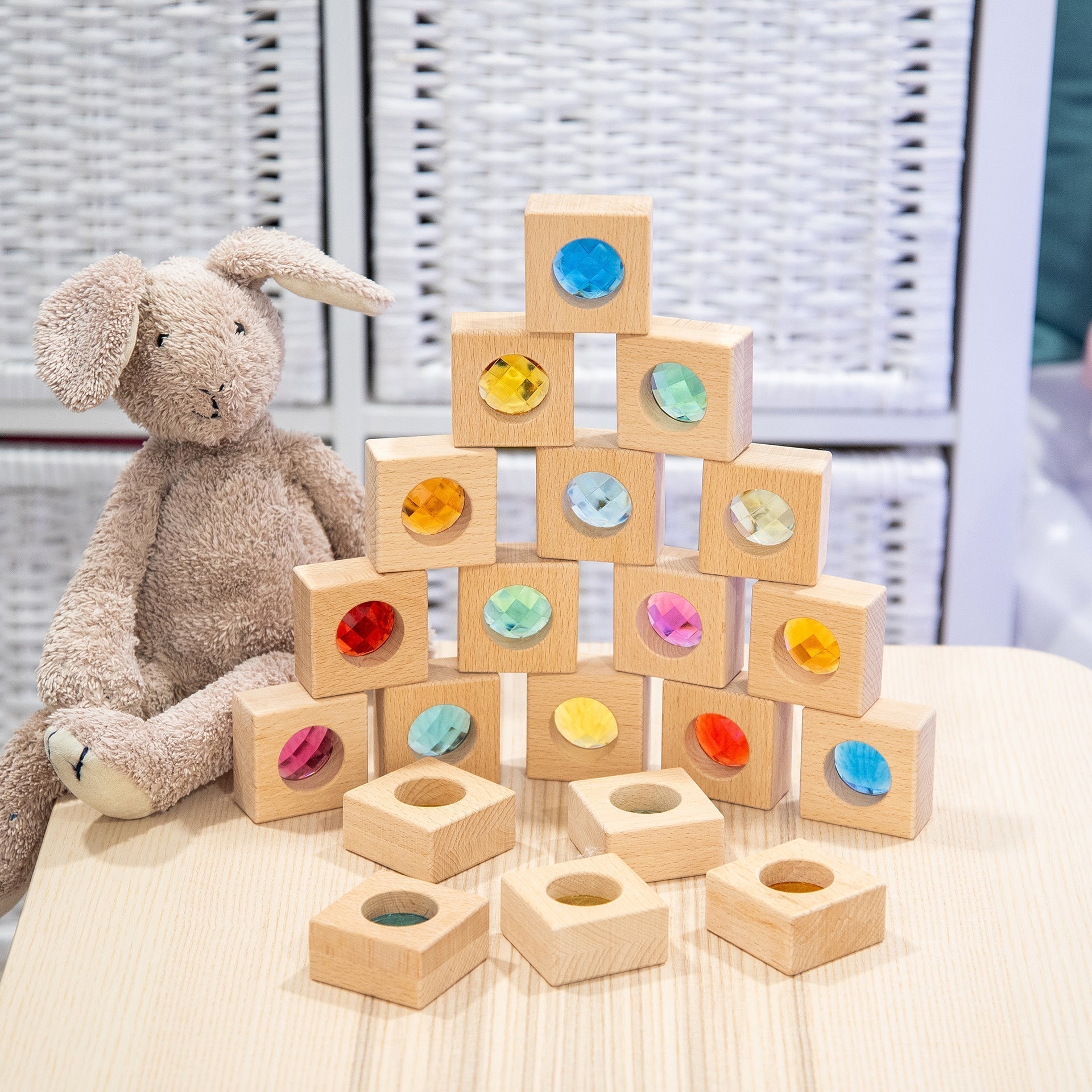 Stack of wooden blocks with colorful recessed shapes next to a plush toy rabbit on a light surface.