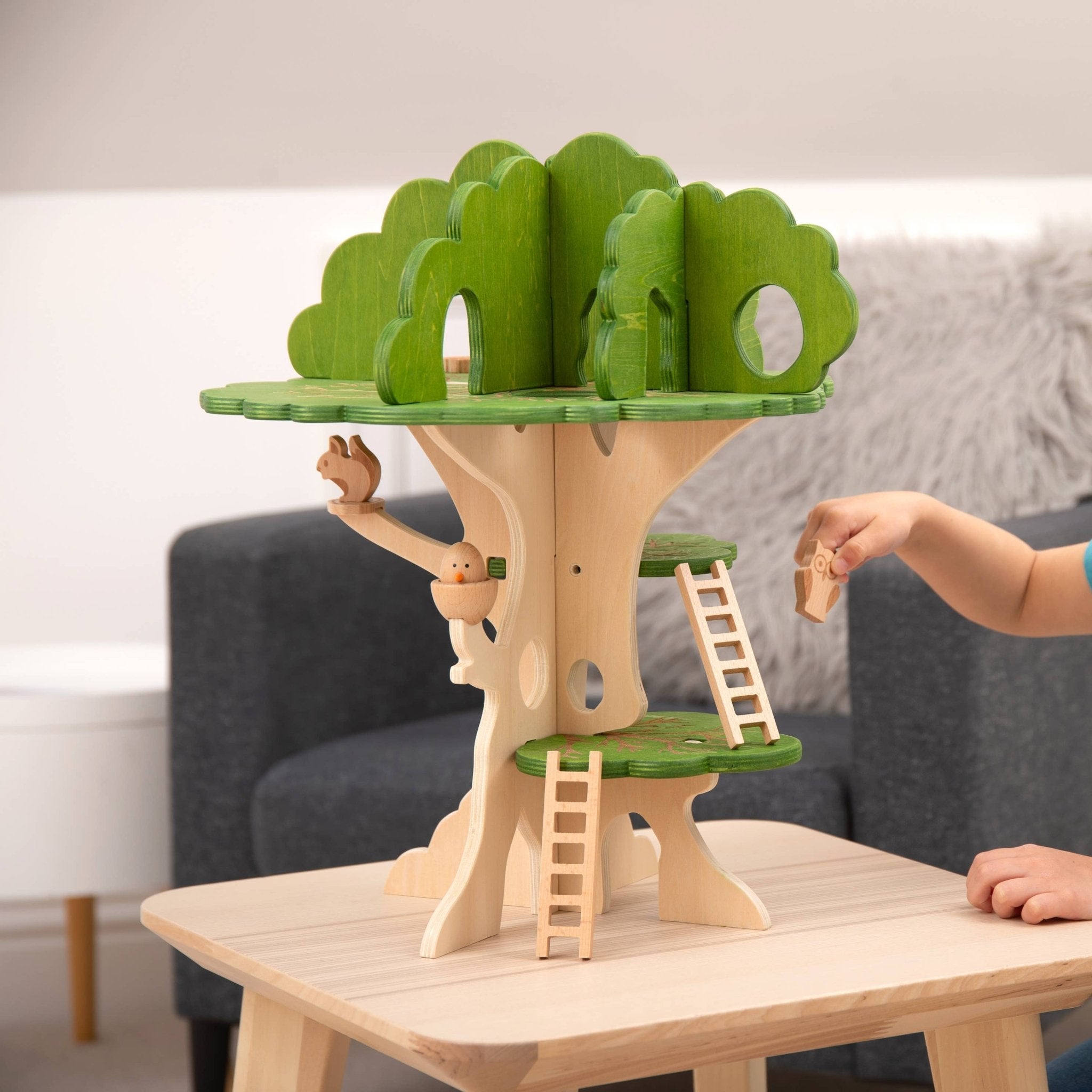 Wooden treehouse toy with green roof and ladders on a table, child's hand reaching out.